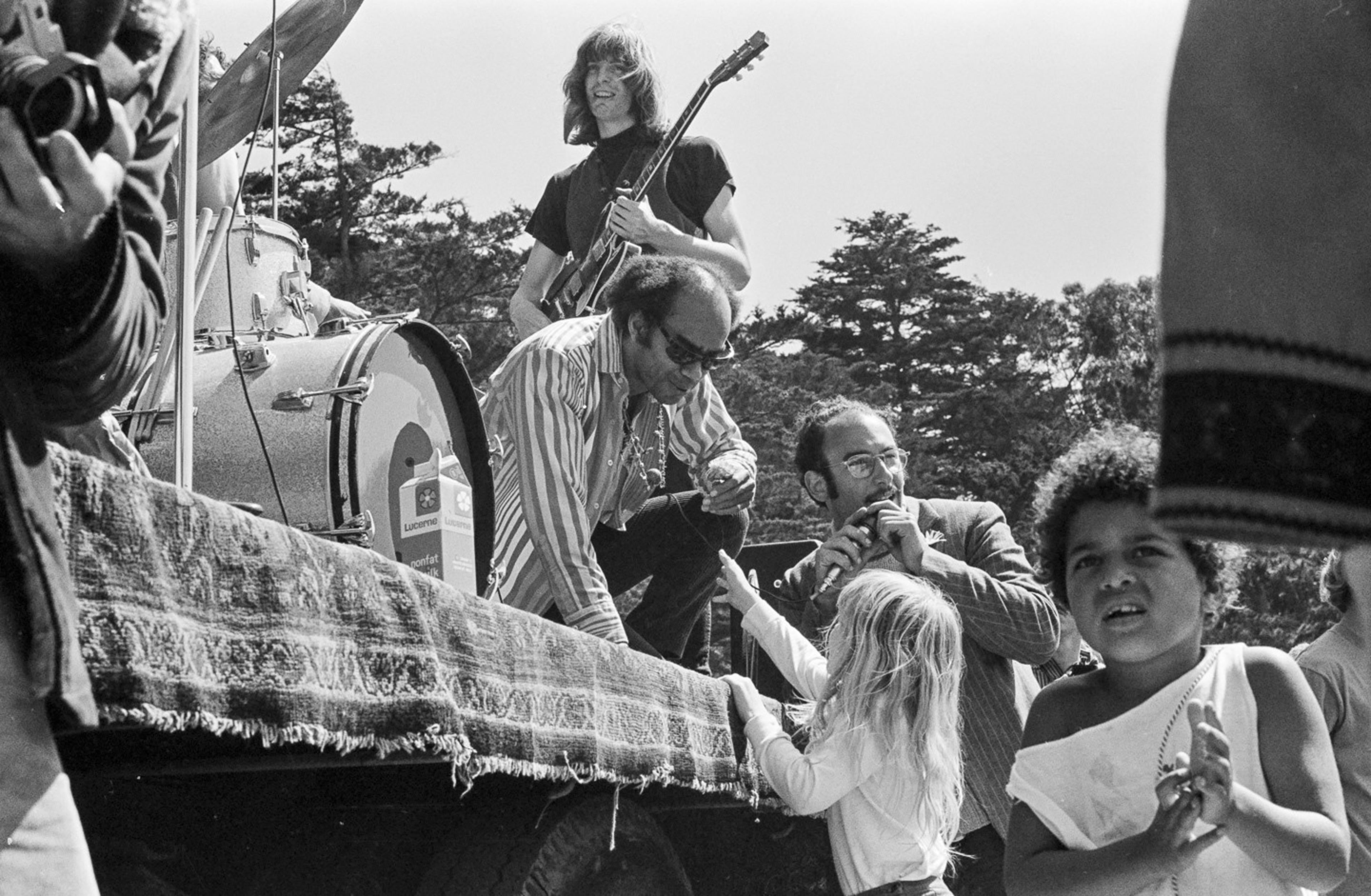 A group of musicians is performing outdoors; a guitarist stands while others kneel, talking to a young child. The scene is lively, with people in motion.