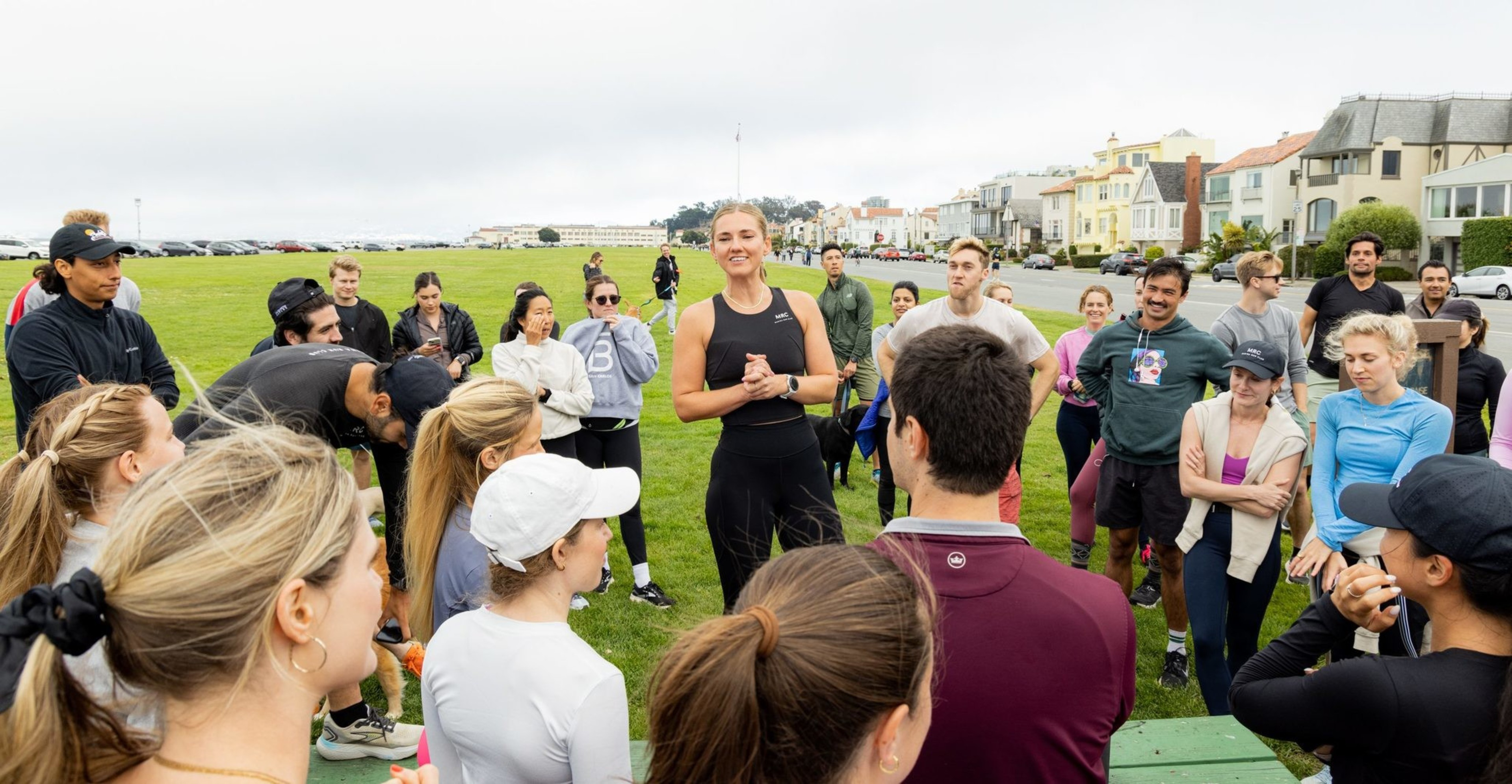 A group of people is gathered on a grassy area, with a woman in the center speaking to them. They appear to be dressed for outdoor activities, with houses visible in the background.