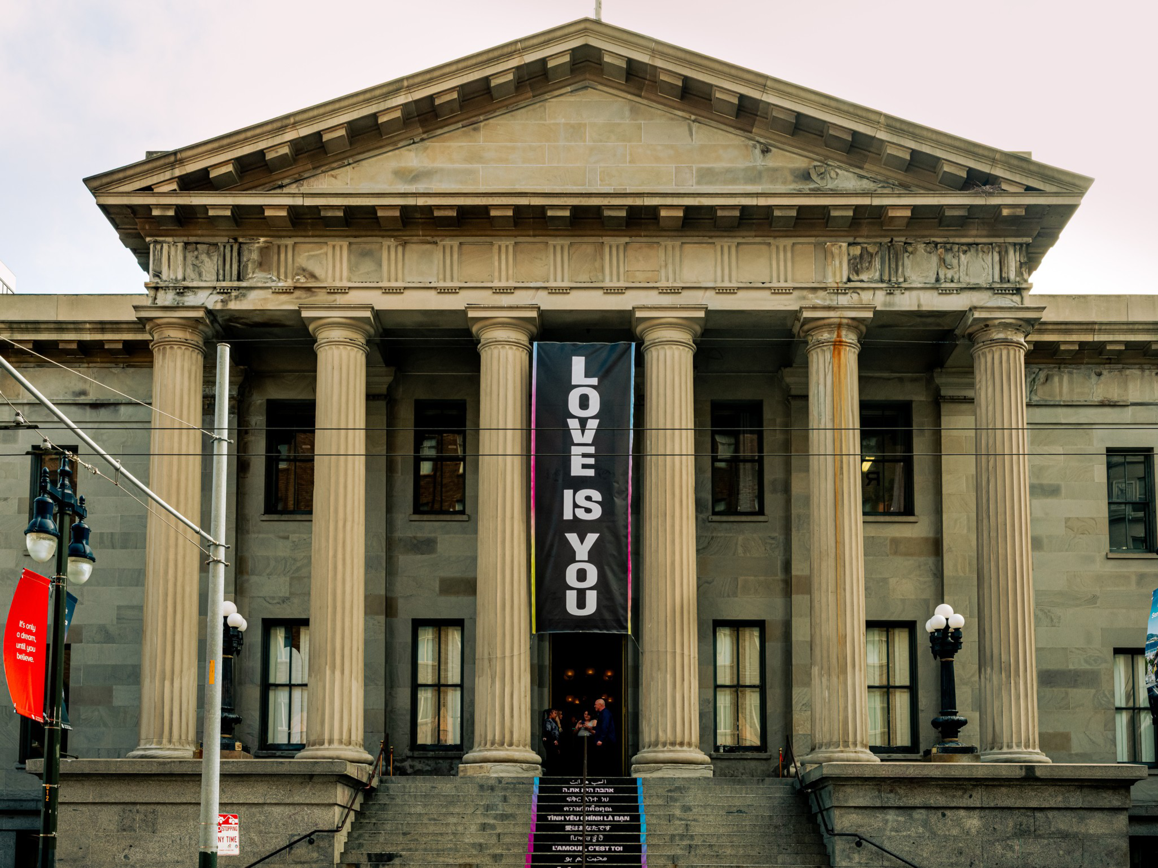 A grand stone building with six tall columns displays a vertical banner that reads “LOVE IS YOU” above the entrance steps.