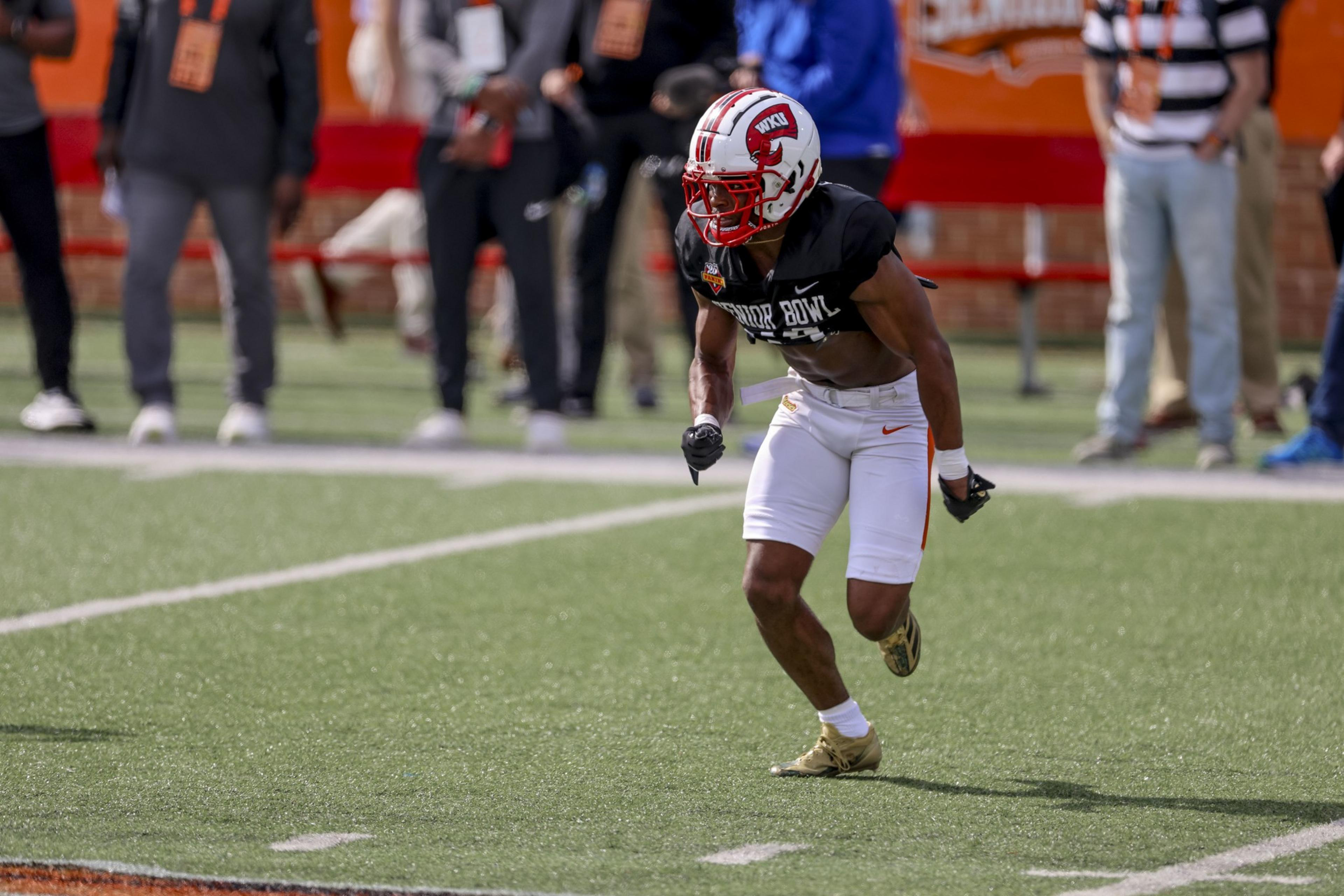 A football player wearing a red and white helmet and a black jersey is crouched in motion on a field, surrounded by onlookers in casual attire.