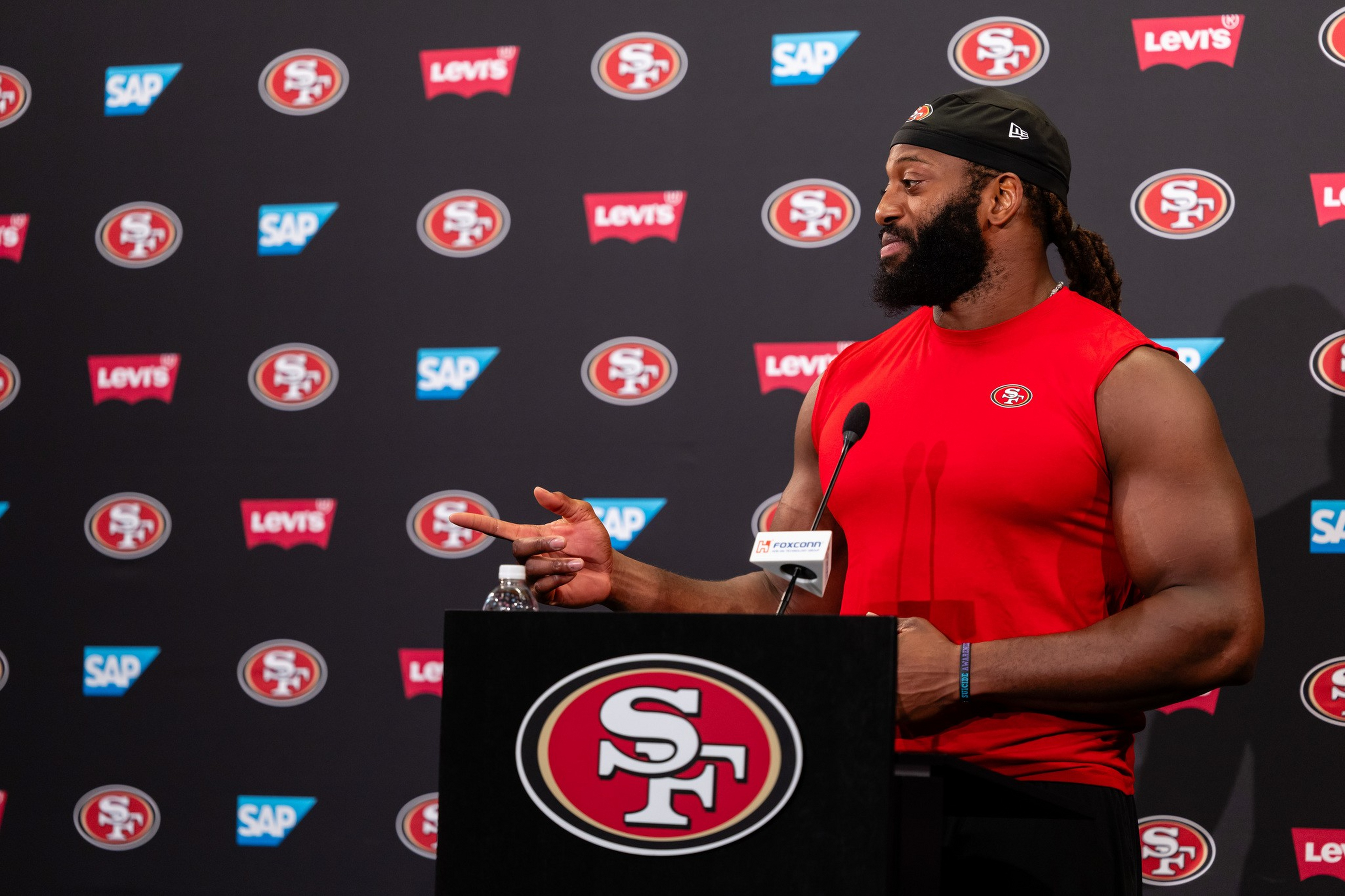 A man in a red sleeveless shirt with an SF logo stands at a podium, pointing to his left, in front of a backdrop with 49ers, Levi's, and SAP logos.