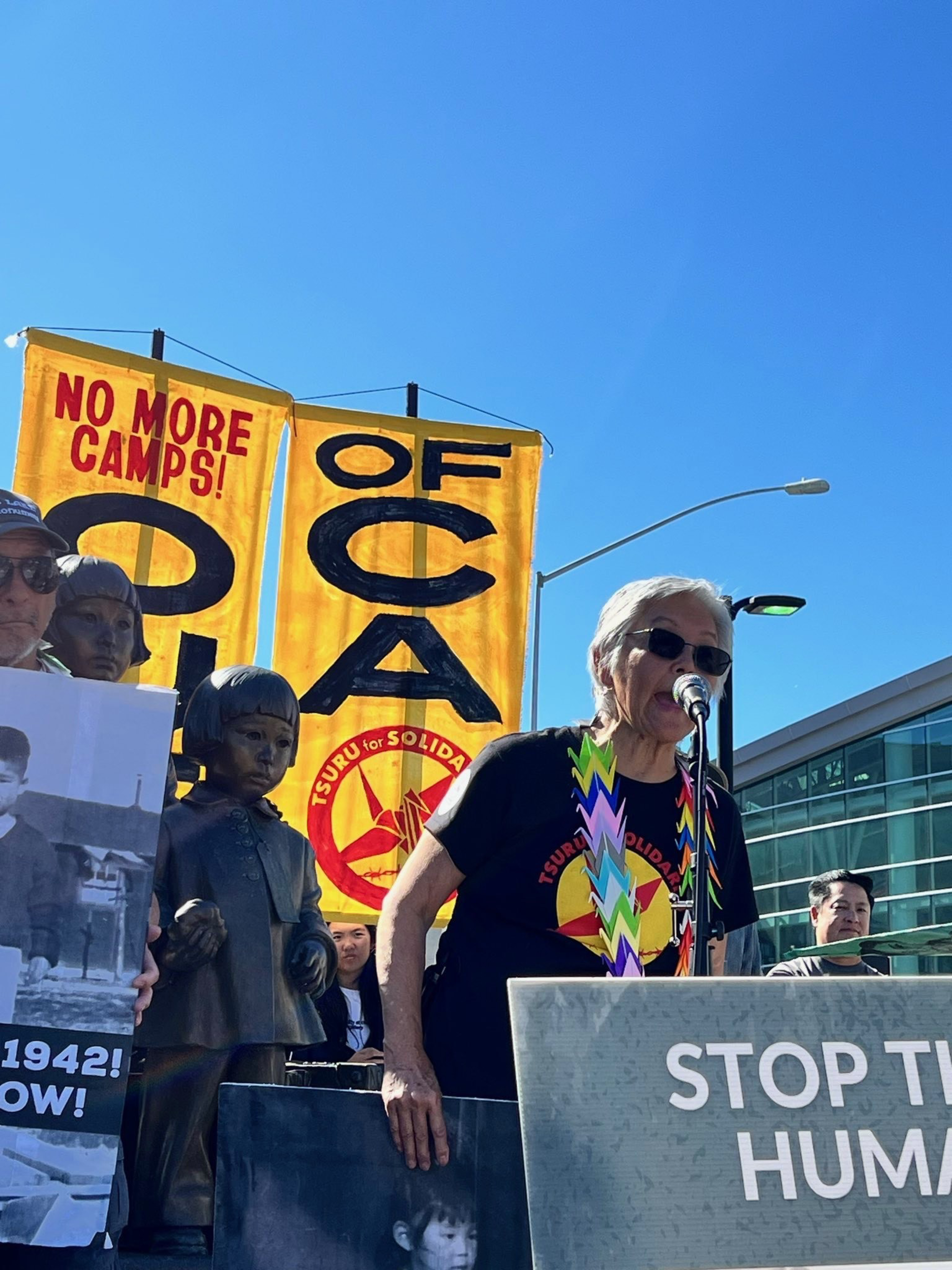 An elderly person speaks into a microphone at a protest with signs reading “NO MORE CAMPS!” and “STOP THE HUMAN,” standing near statues of children.