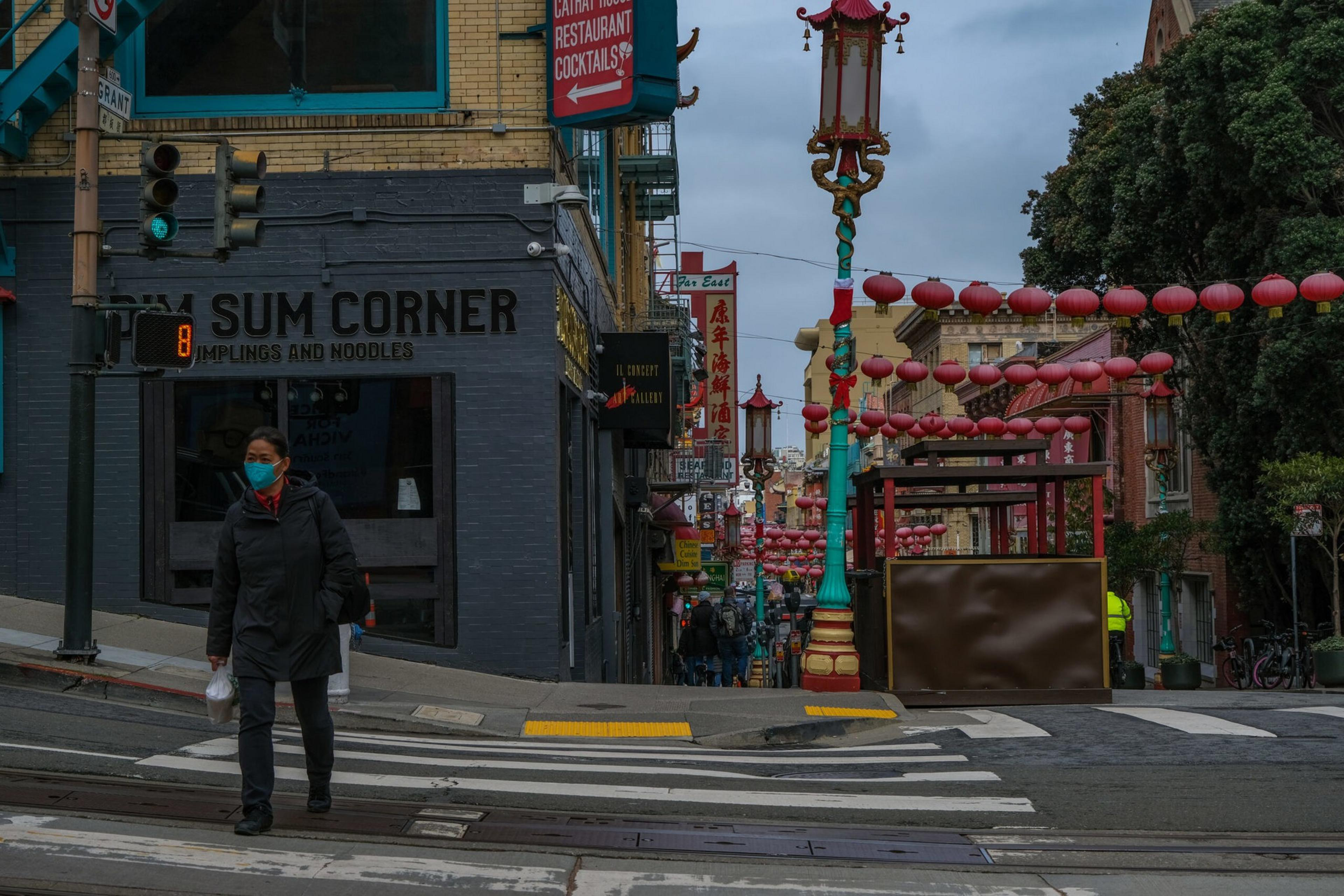 A person wearing a mask crosses a street near a shop called "Dim Sum Corner" in a Chinatown area decorated with red lanterns and colorful street lamps.