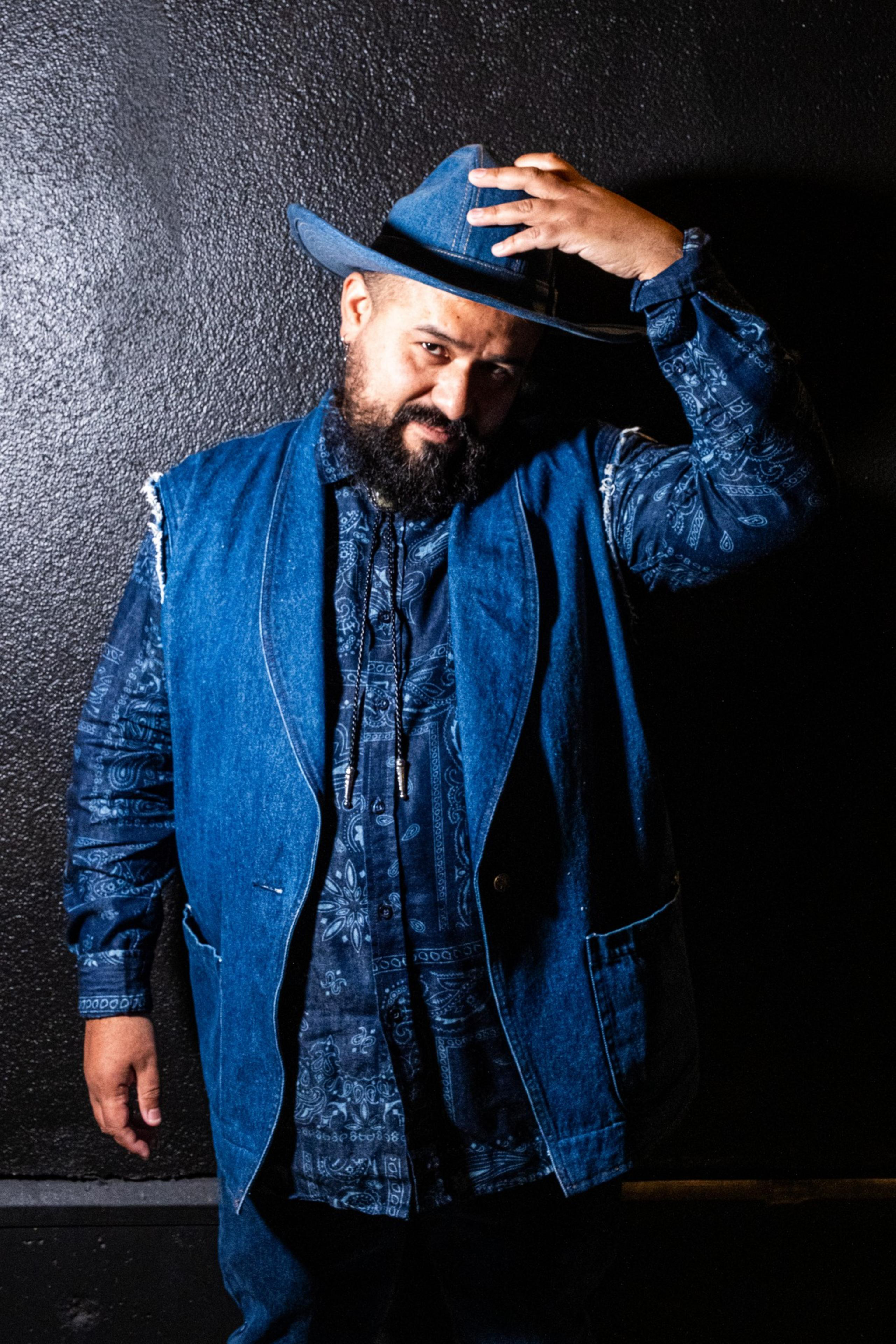 A man stands against a dark background, wearing a denim hat and vest over a patterned shirt. He has a beard and is adjusting his hat with one hand.