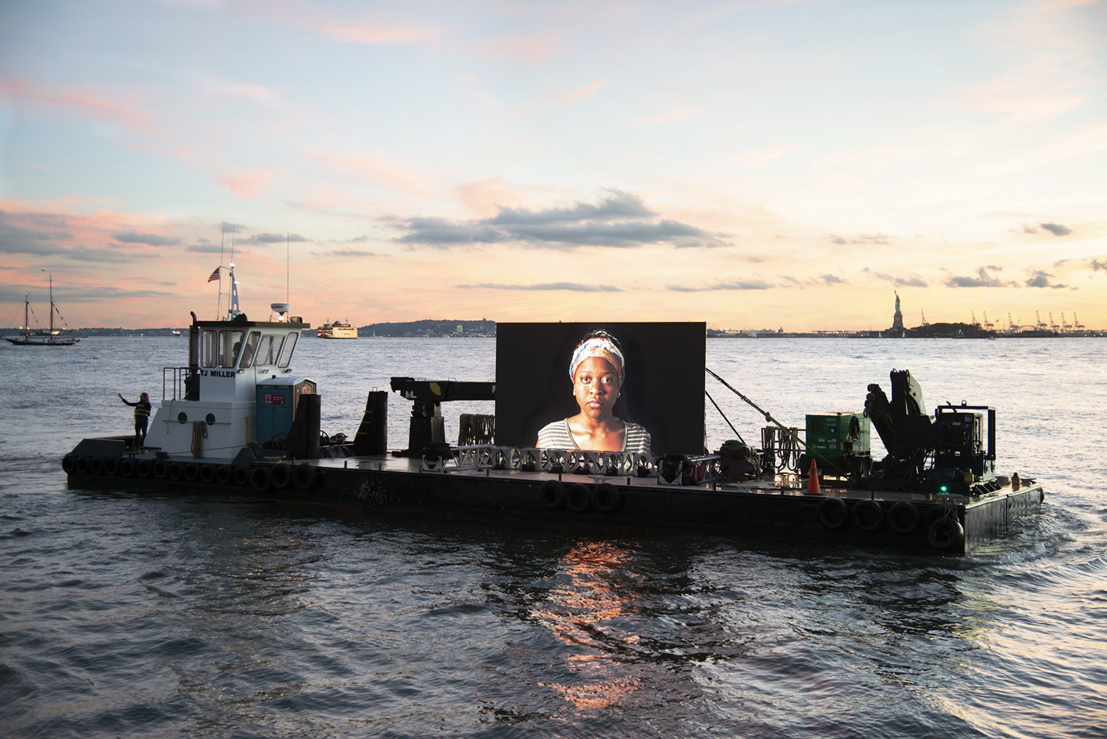 A barge floats on calm water at sunset, displaying a large illuminated portrait of a woman with a headscarf, with the Statue of Liberty visible in the distance.