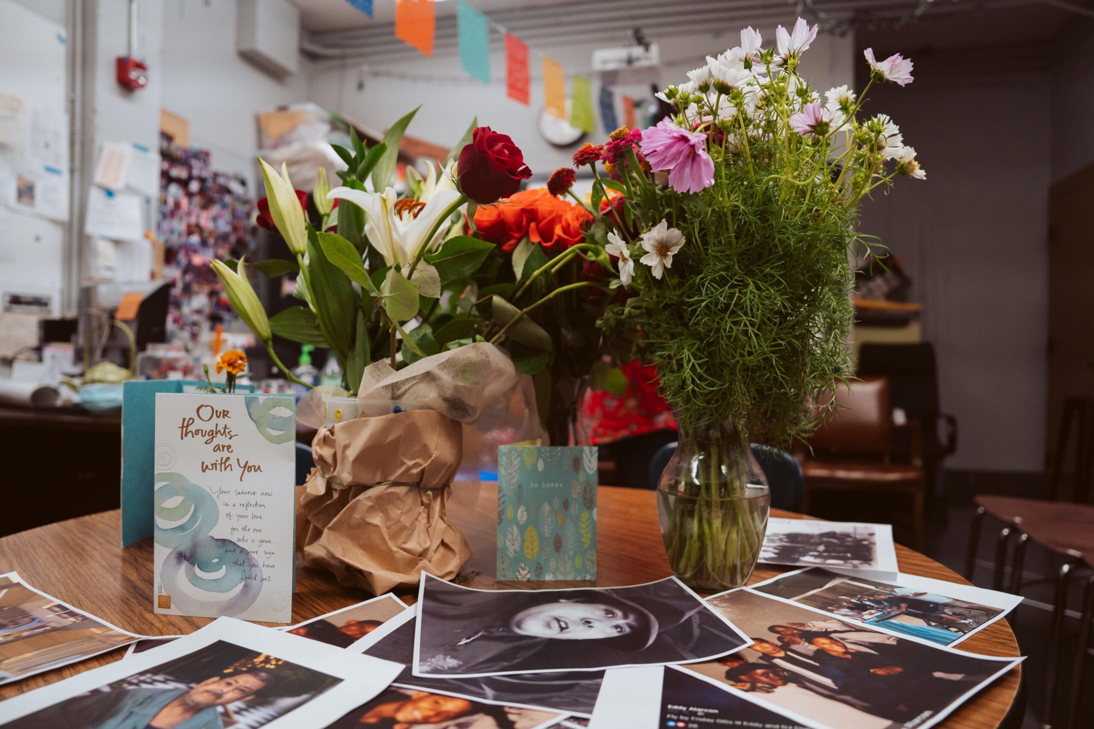 A table holds bouquets of flowers, sympathy cards, and numerous printed photos of people, suggesting a memorial or tribute setting.