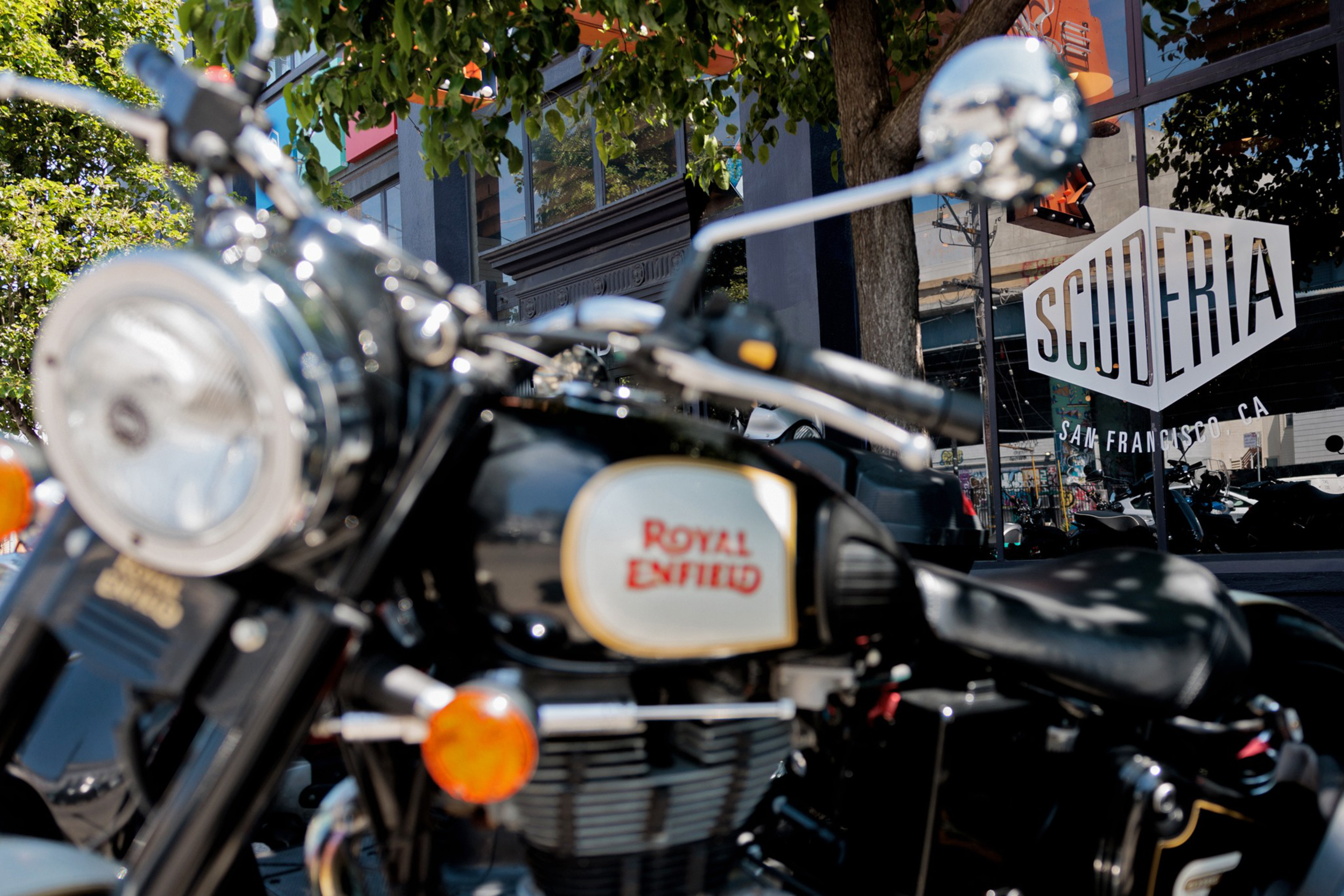 A black Royal Enfield motorcycle is parked outdoors near a tree, in front of a building with a glass window displaying a "SCUDERIA SAN FRANCISCO, CA" sign.