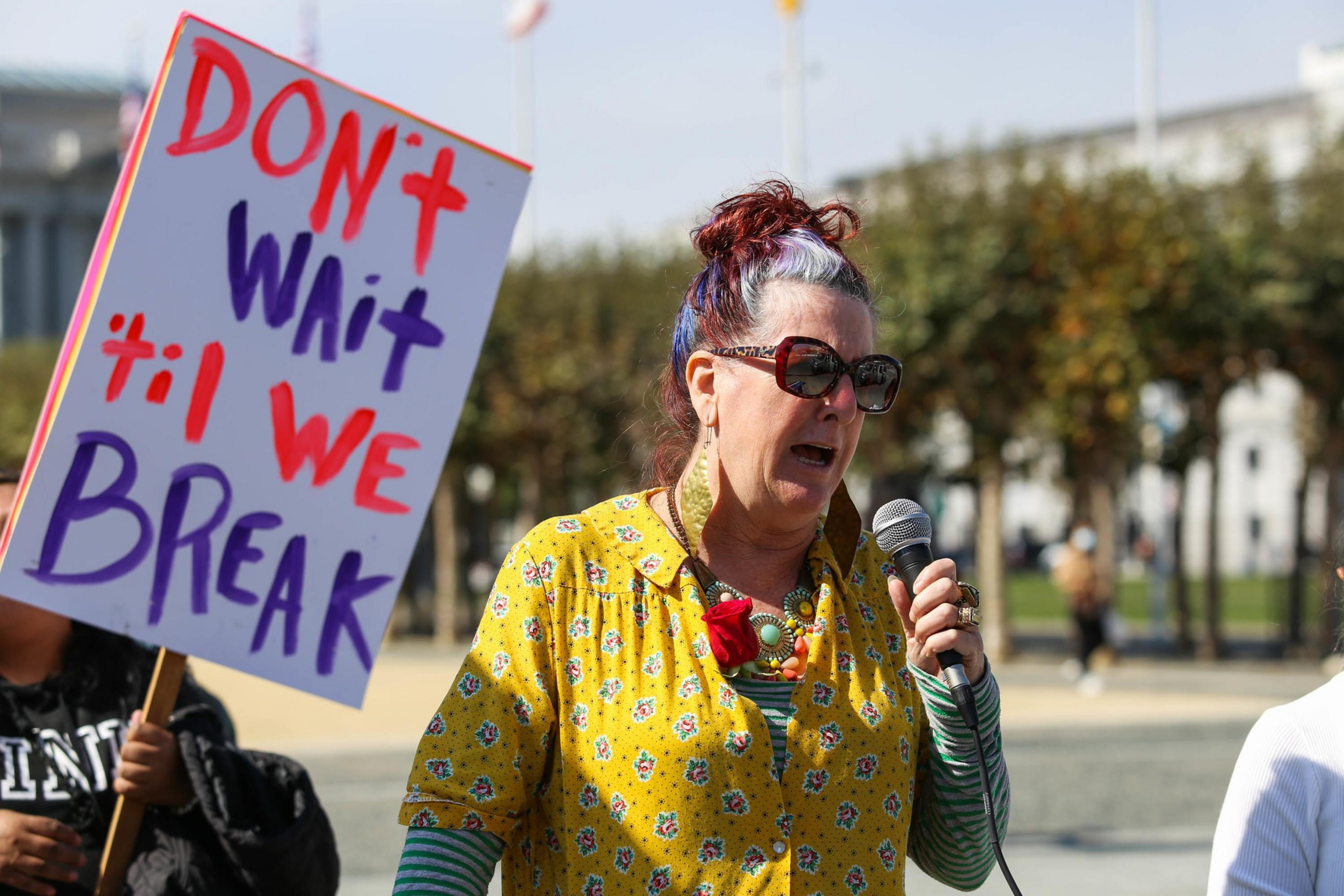 A woman with sunglasses and colorful hair speaks into a microphone while another holds a sign reading, "DON'T wait 'til WE BREAK."