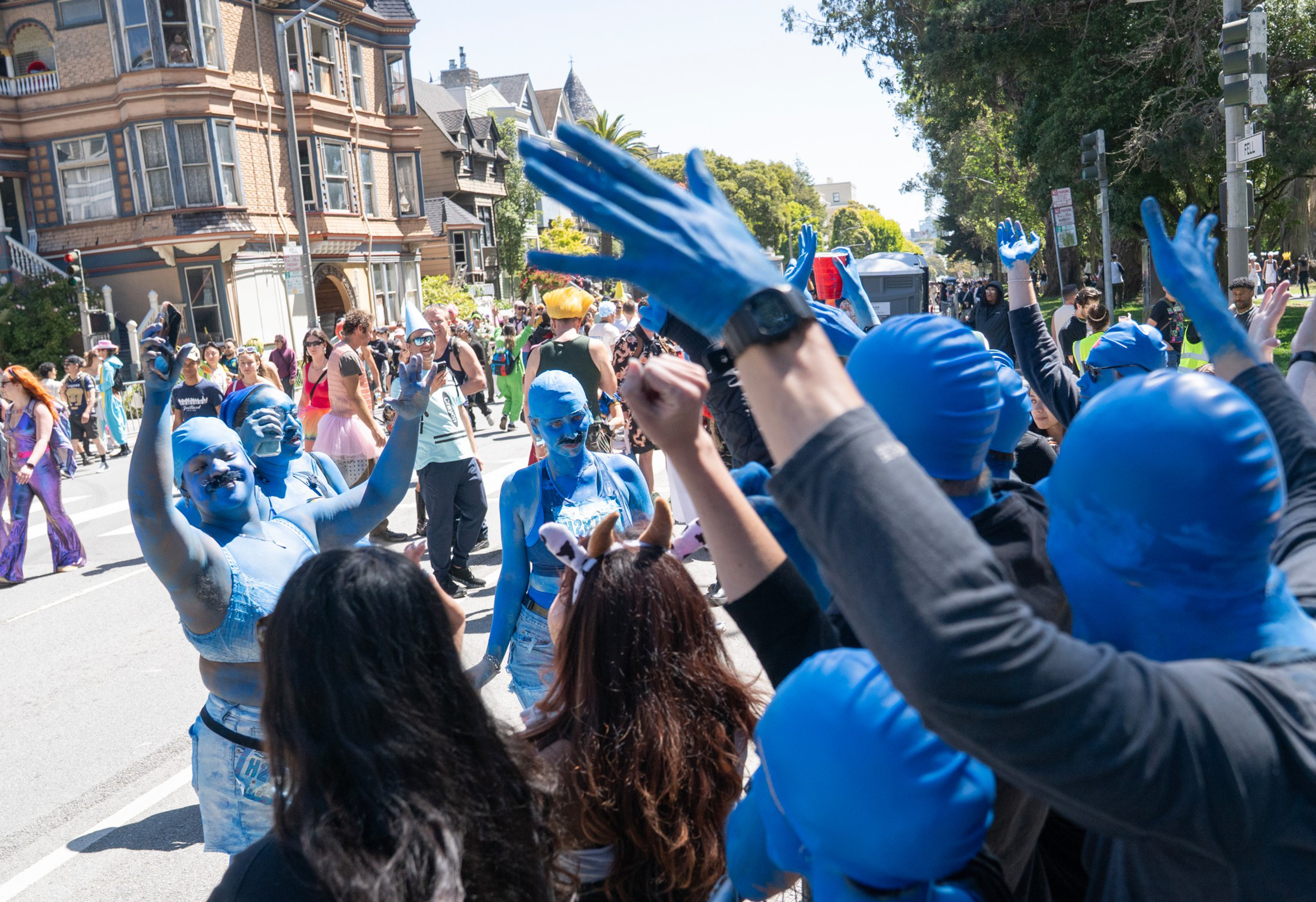 People in blue full-body suits and painted blue face and bodies raise their hands and celebrate on a sunny street during a festive outdoor event.