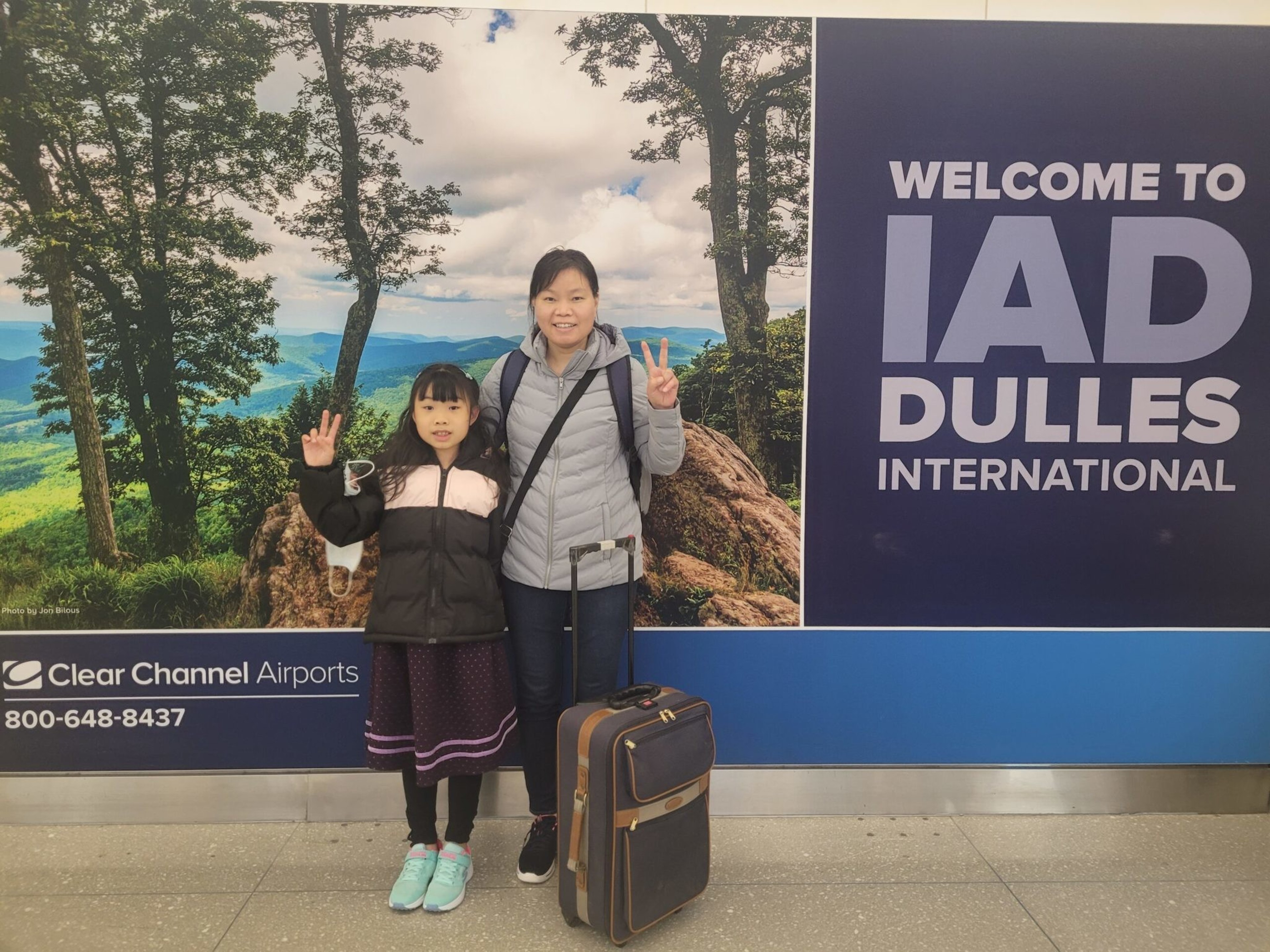 A woman and child stand with peace signs in front of a "Welcome to IAD Dulles International" sign, with a suitcase beside them.