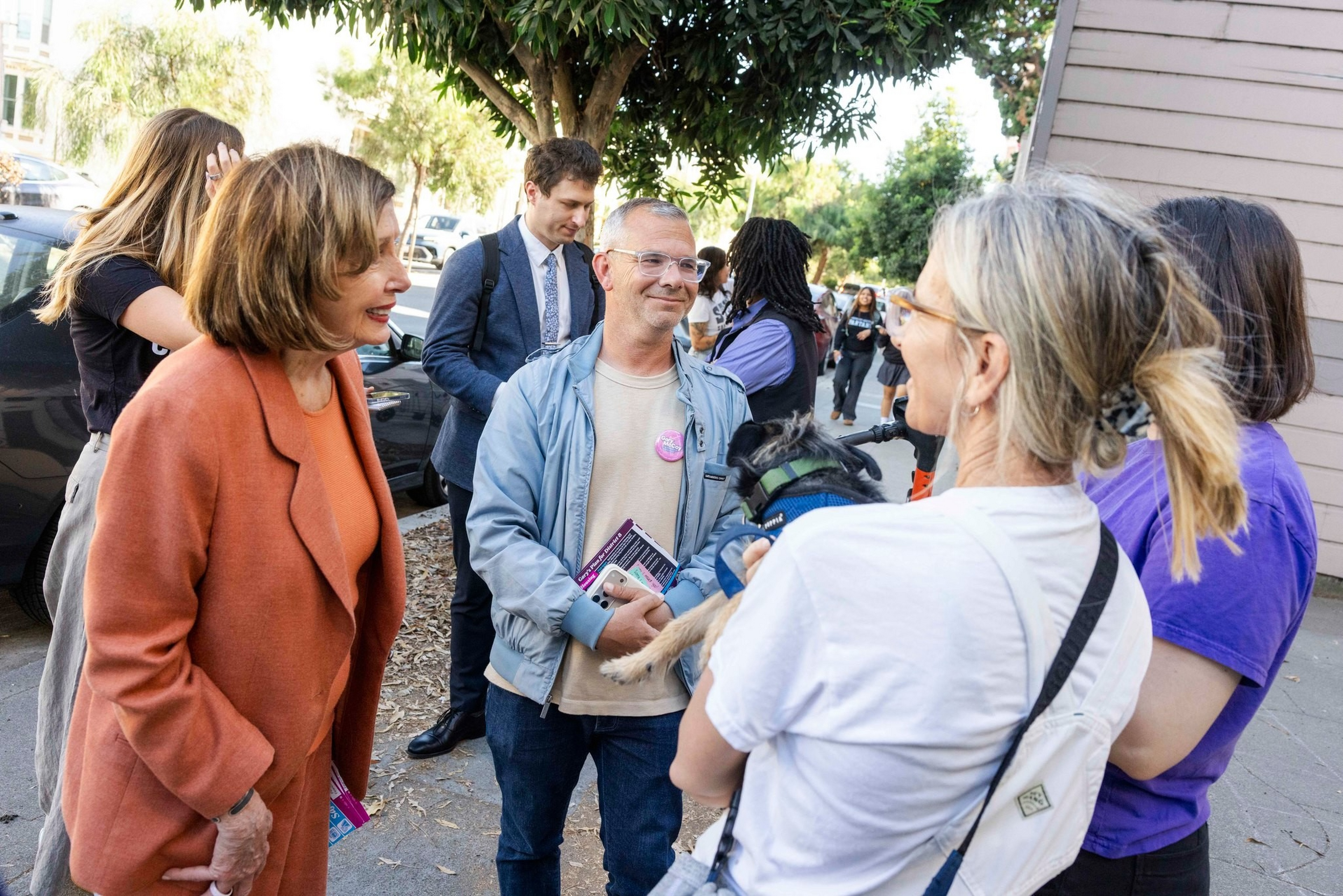A woman in an orange coat talks to a man holding a booklet and a small dog, while two other people stand nearby on a sidewalk by a tree.