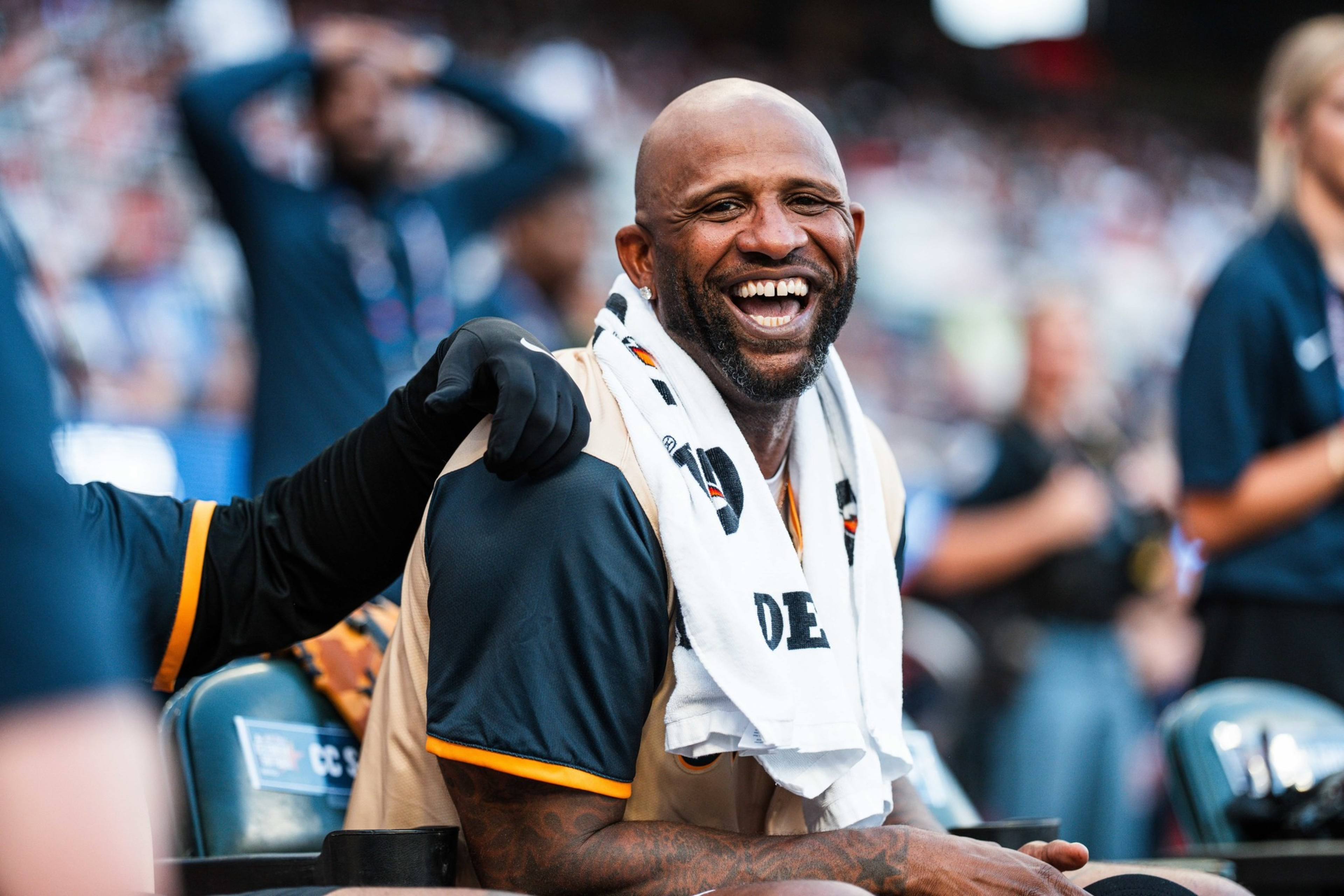 A smiling man sits on a sports bench, draped in a towel around his neck. He wears a dark shirt, and others are blurred in the background at a crowded event.