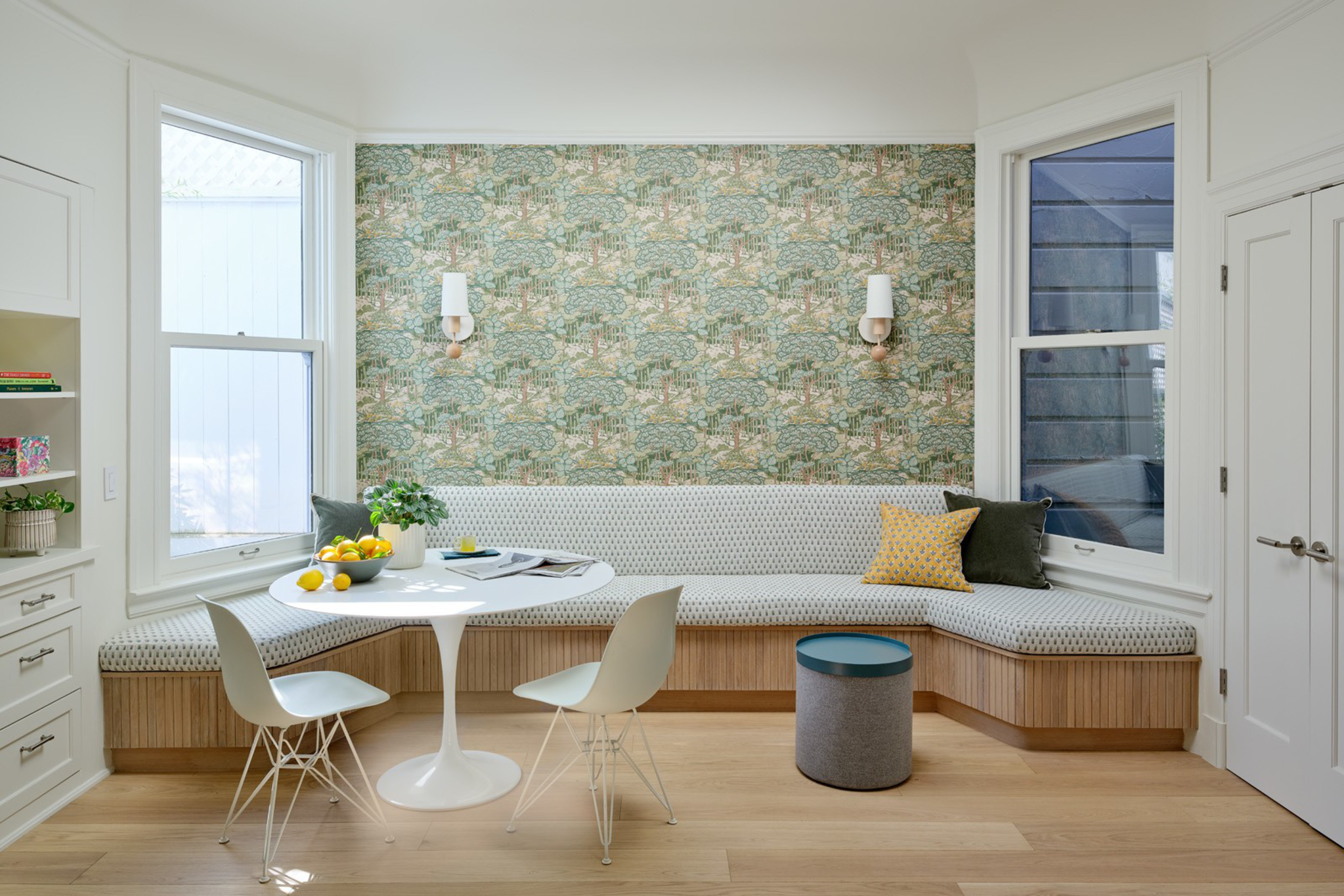 A cozy breakfast nook with patterned wallpaper, a cushioned bench with throw pillows, a round white table, two white chairs, a small blue stool, and large windows.
