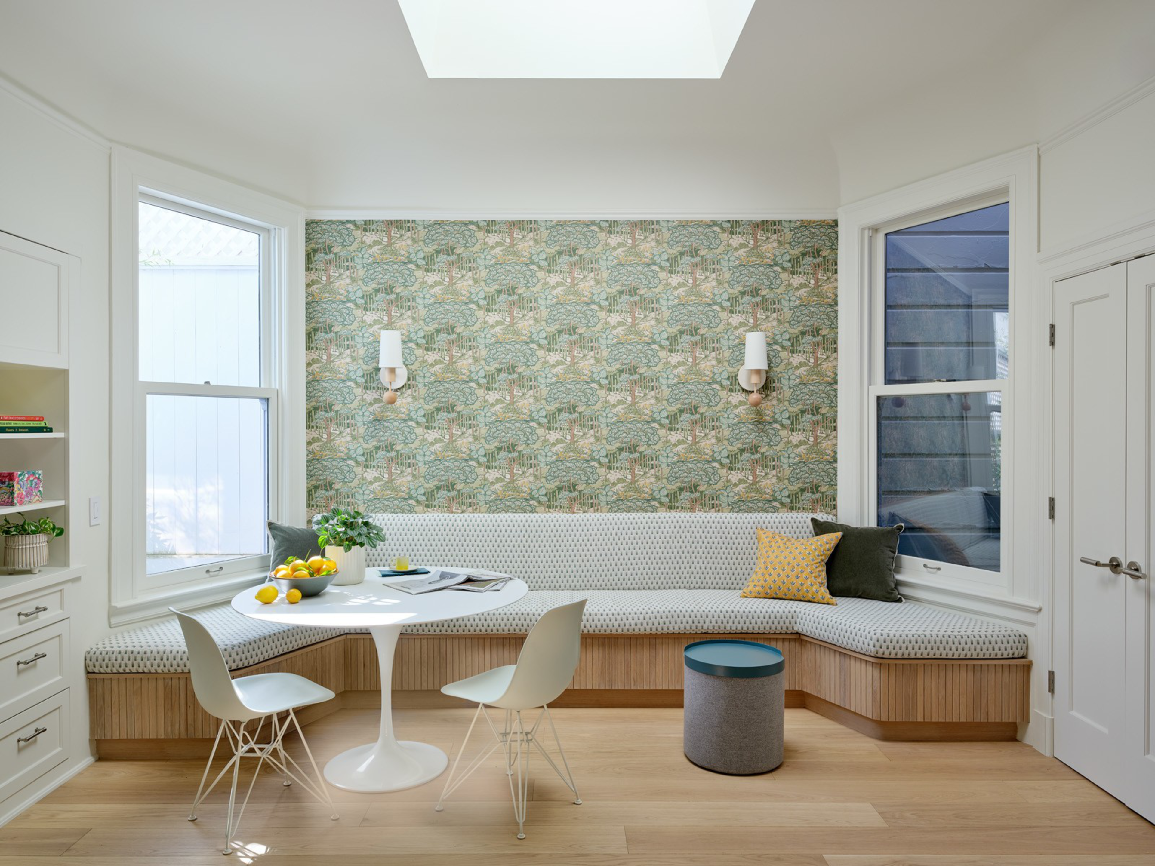A cozy breakfast nook with patterned wallpaper, a cushioned bench with throw pillows, a round white table, two white chairs, a small blue stool, and large windows.
