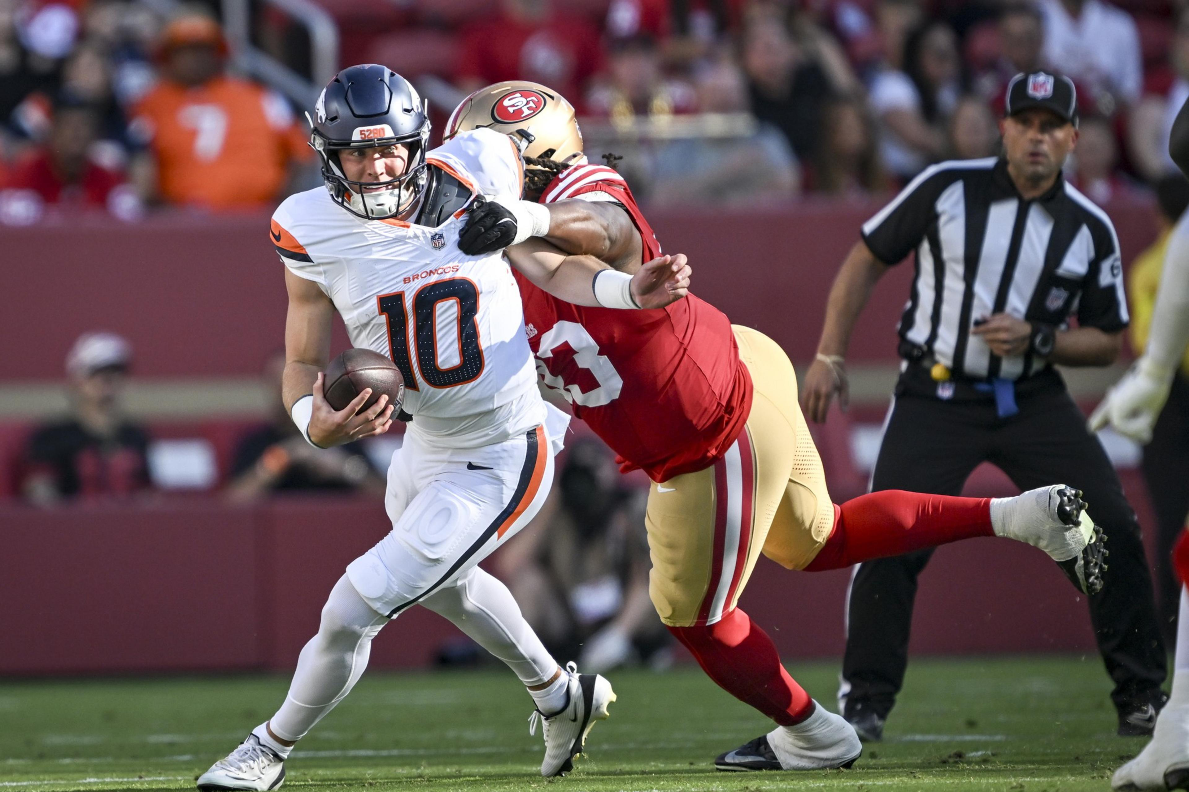 A football player in white and orange is running with the ball while a player in red and gold tries to tackle him from behind.
