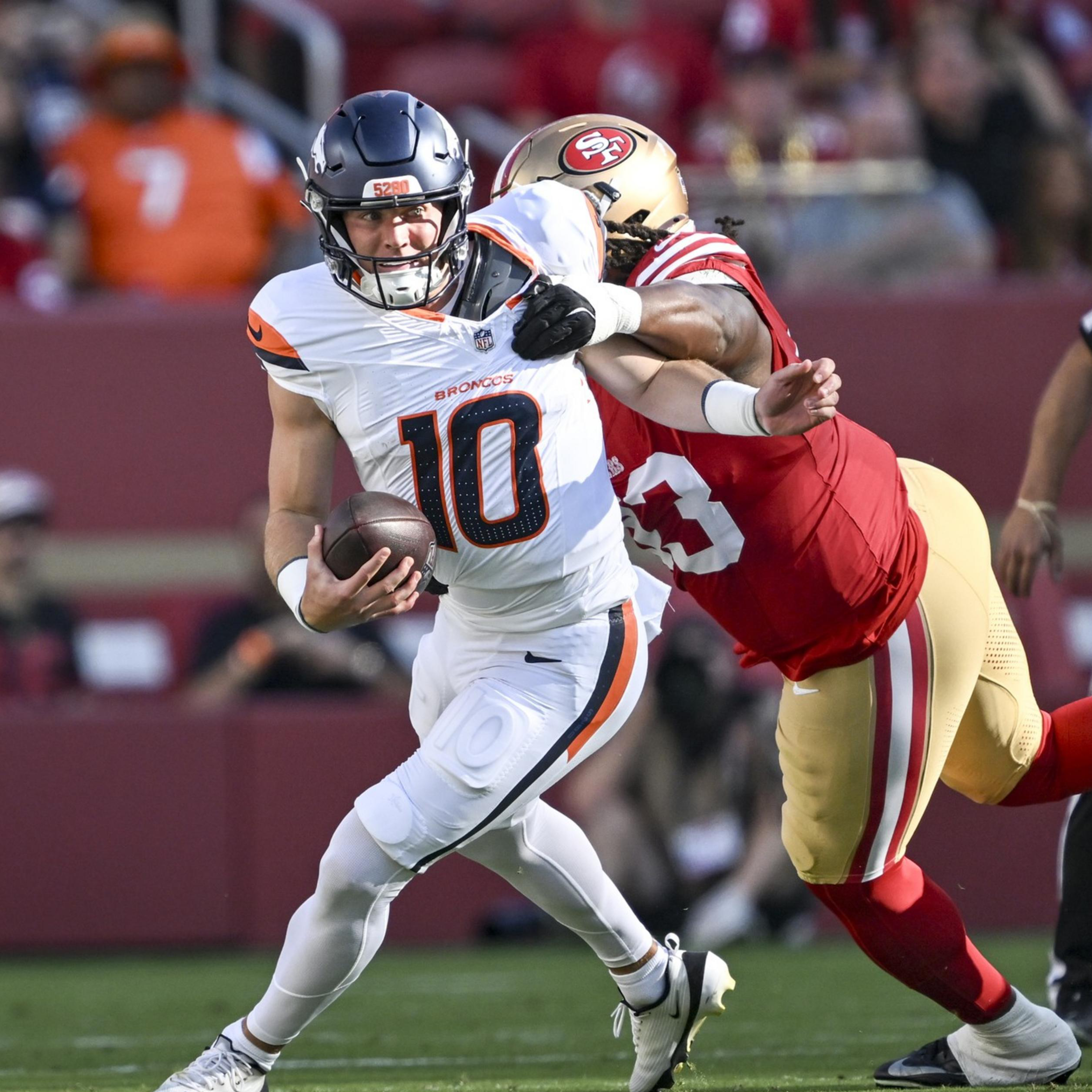 A football player in white and orange is running with the ball while a player in red and gold tries to tackle him from behind.