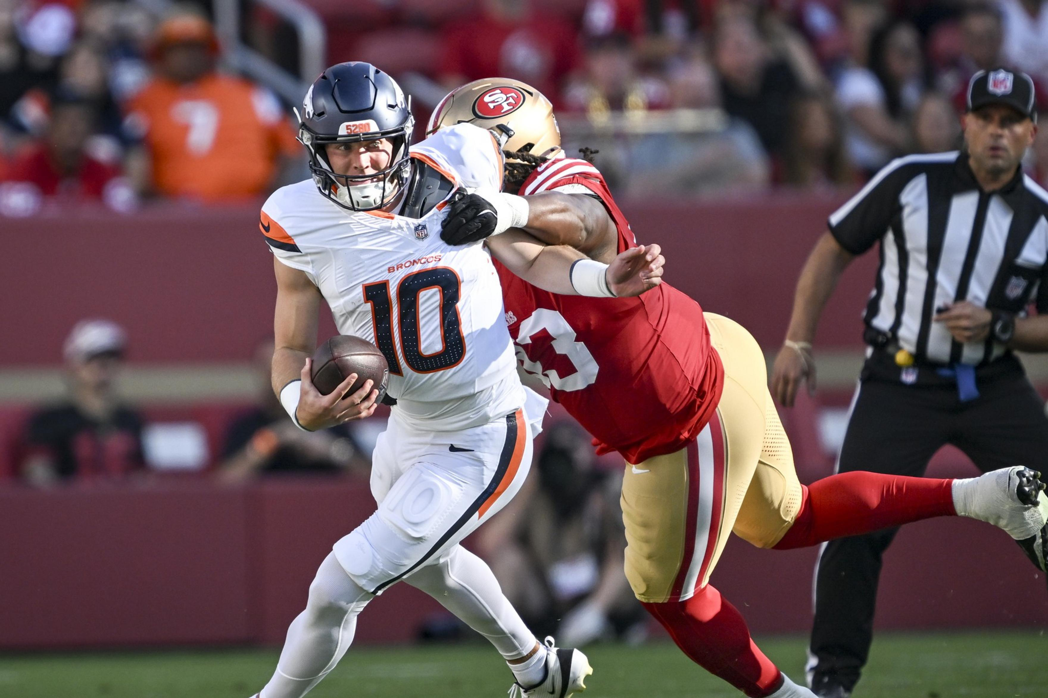 A football player in white and orange is running with the ball while a player in red and gold tries to tackle him from behind.