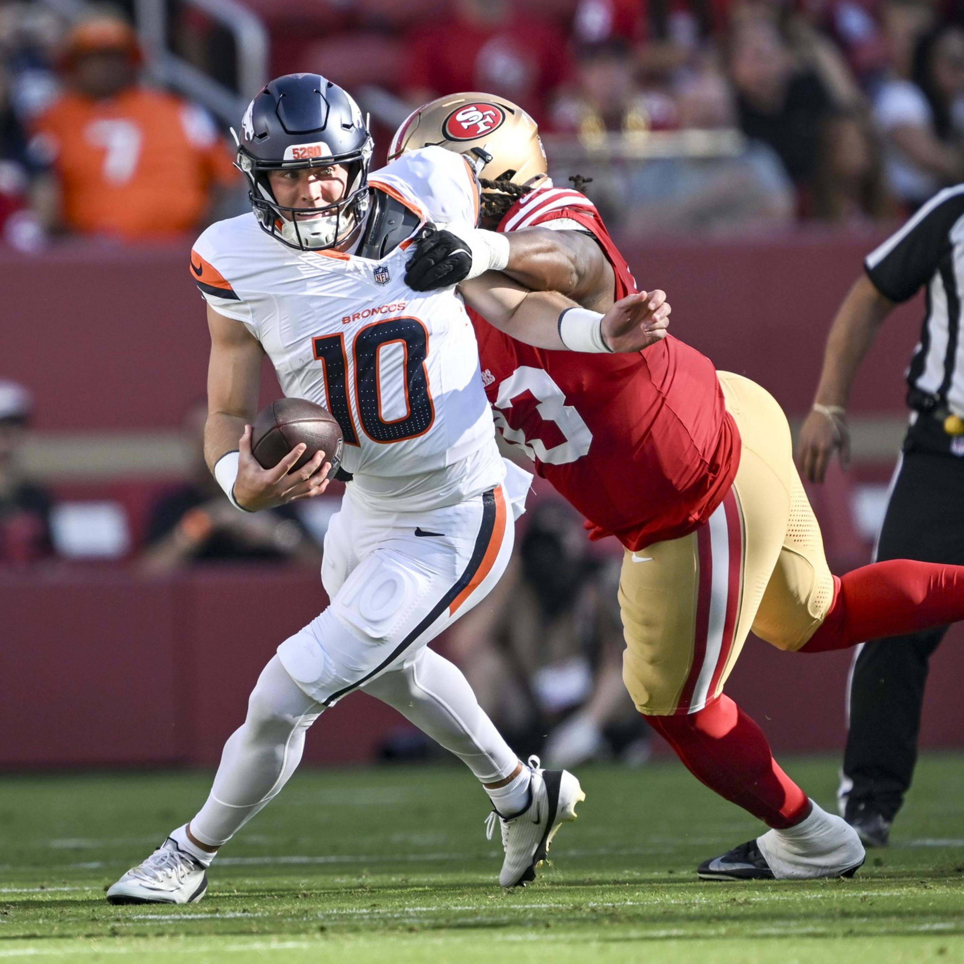 A football player in white and orange is running with the ball while a player in red and gold tries to tackle him from behind.