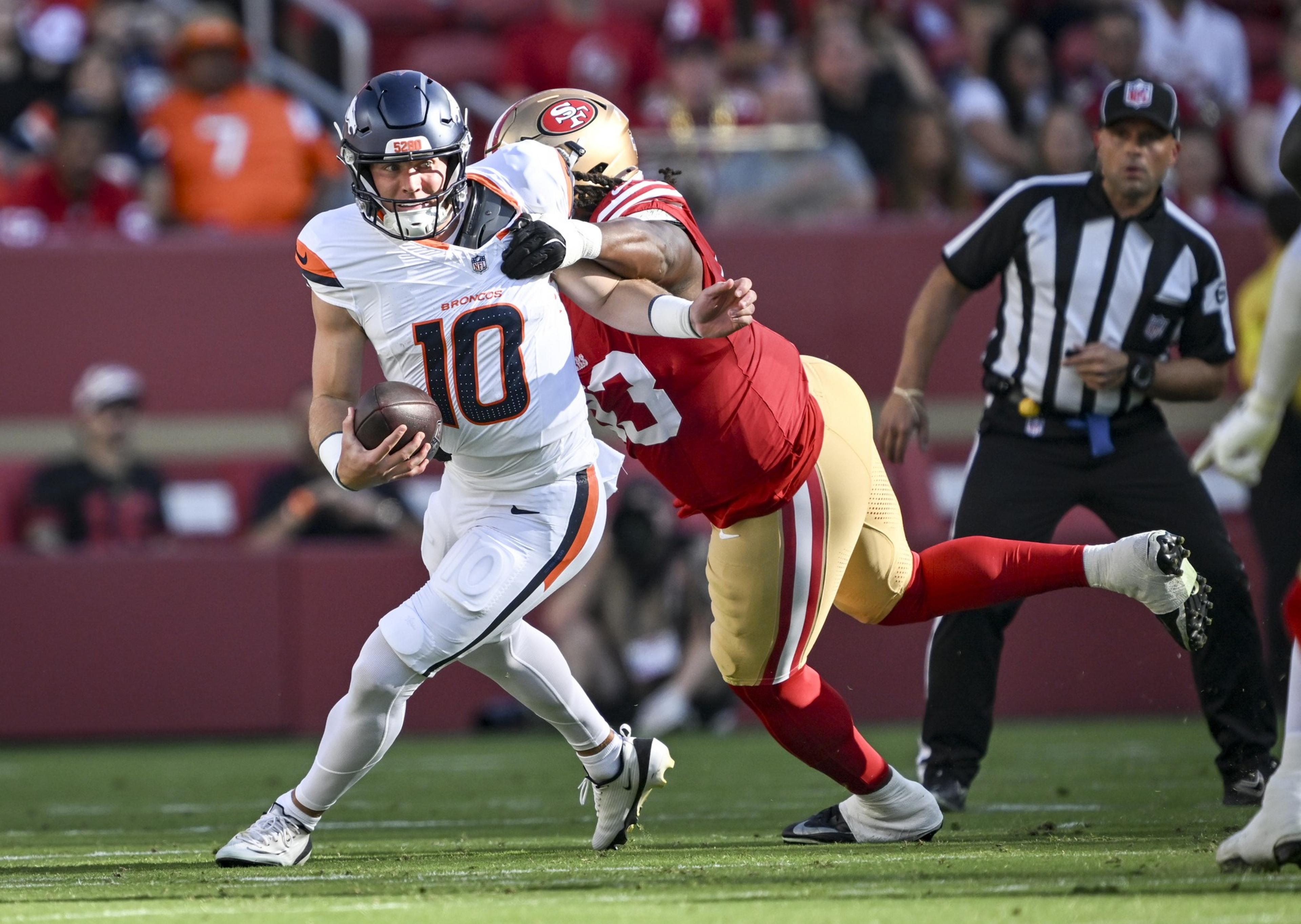 A football player in white and orange is running with the ball while a player in red and gold tries to tackle him from behind.