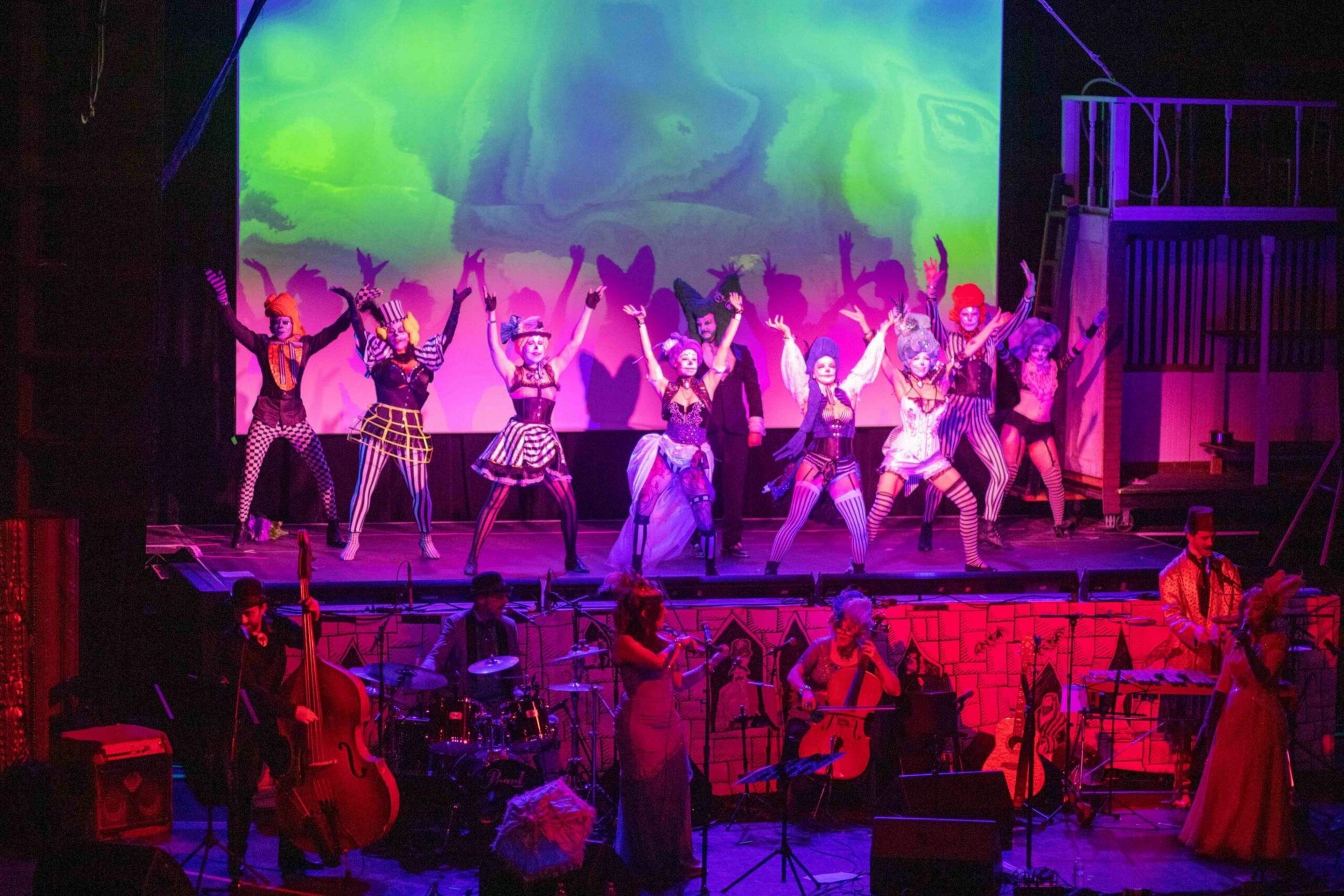 Performers in colorful, theatrical costumes dance with raised arms on stage, while musicians play instruments below under vibrant purple and green lighting.