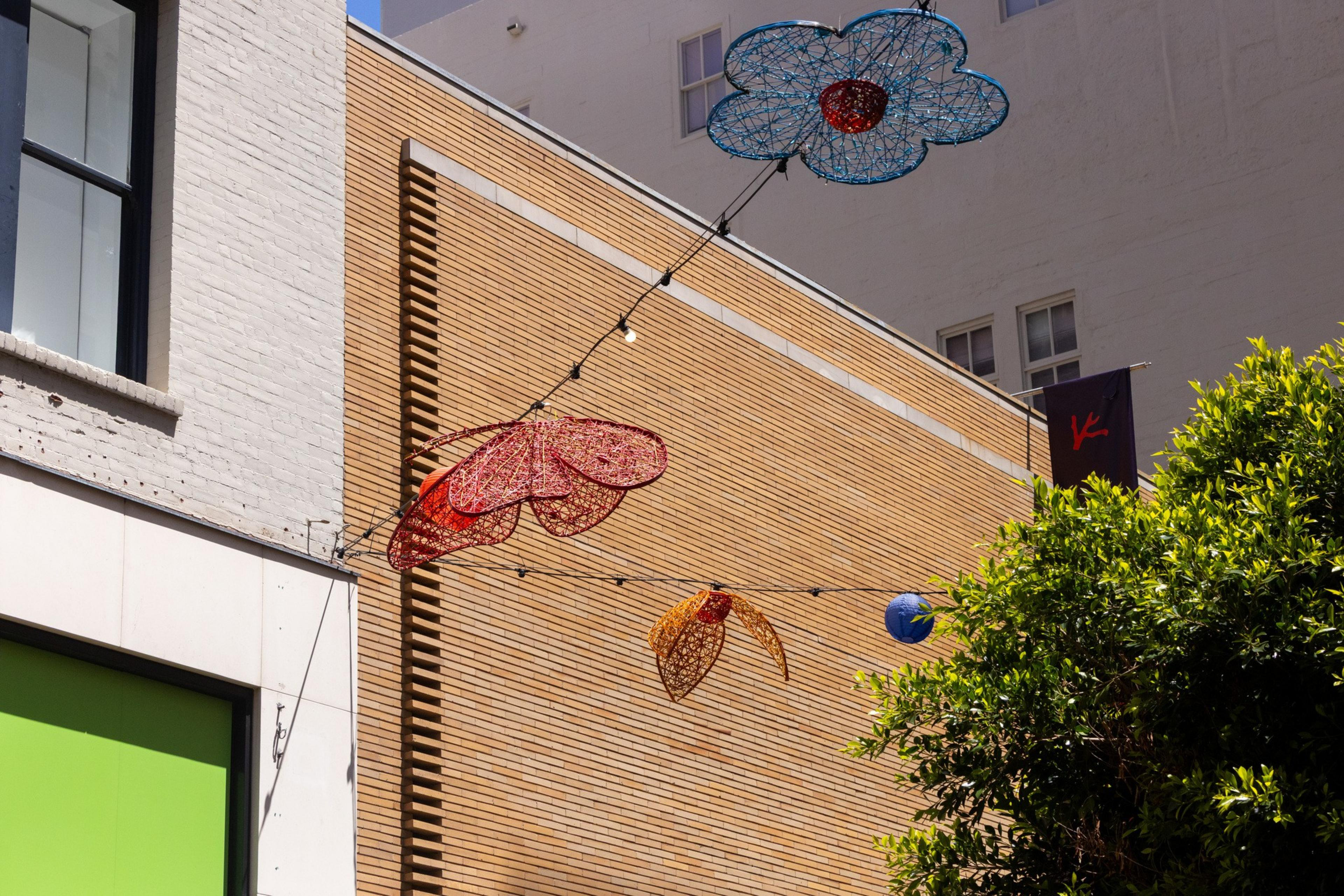 Colorful wire sculptures shaped like flowers hang above a street, set against brick and white walls, with a green window and some trees visible.