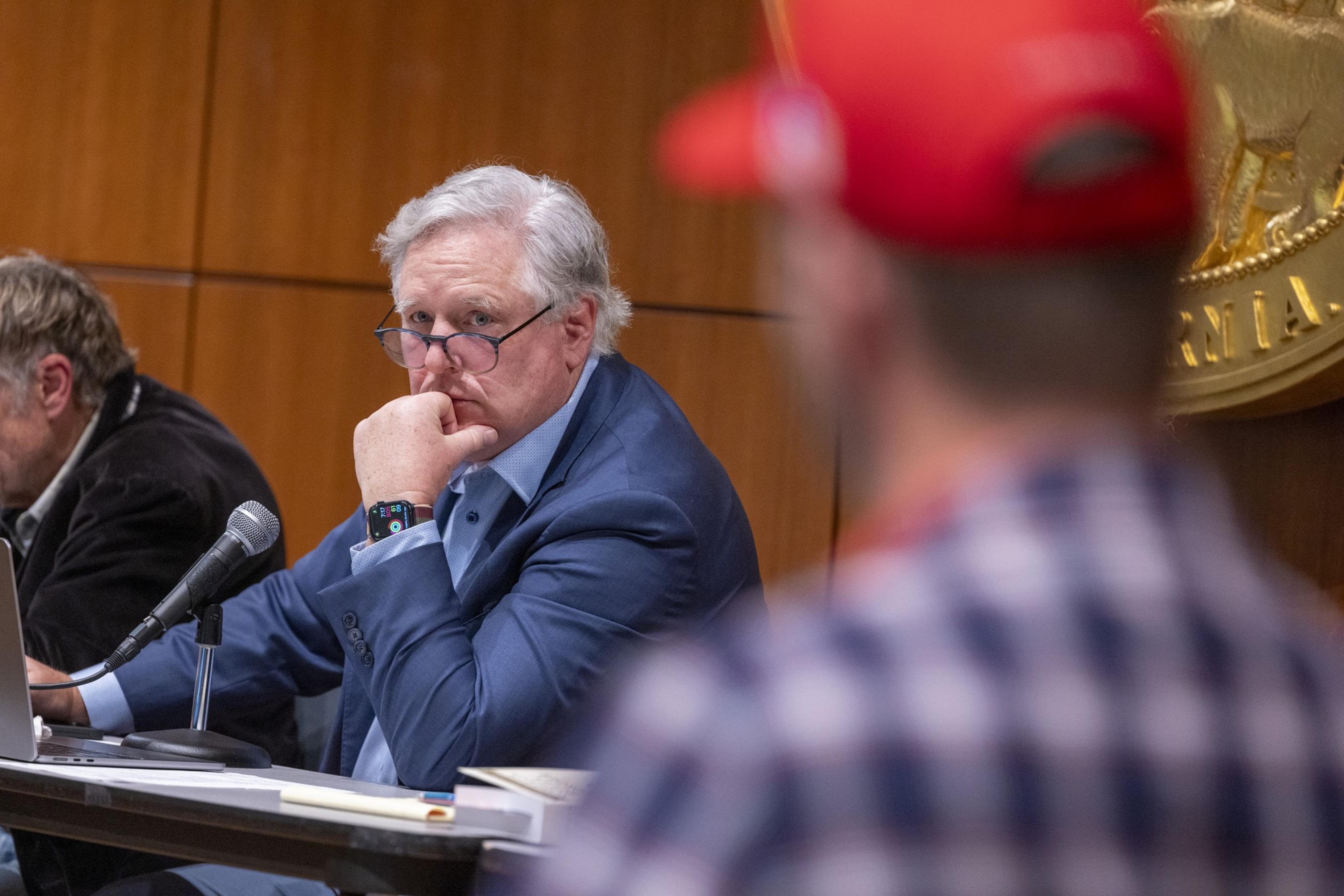 A man in a blue suit and glasses sits at a table, focusing intensely, with his hand on his chin. Another, blurred man in a red cap, is in the foreground.