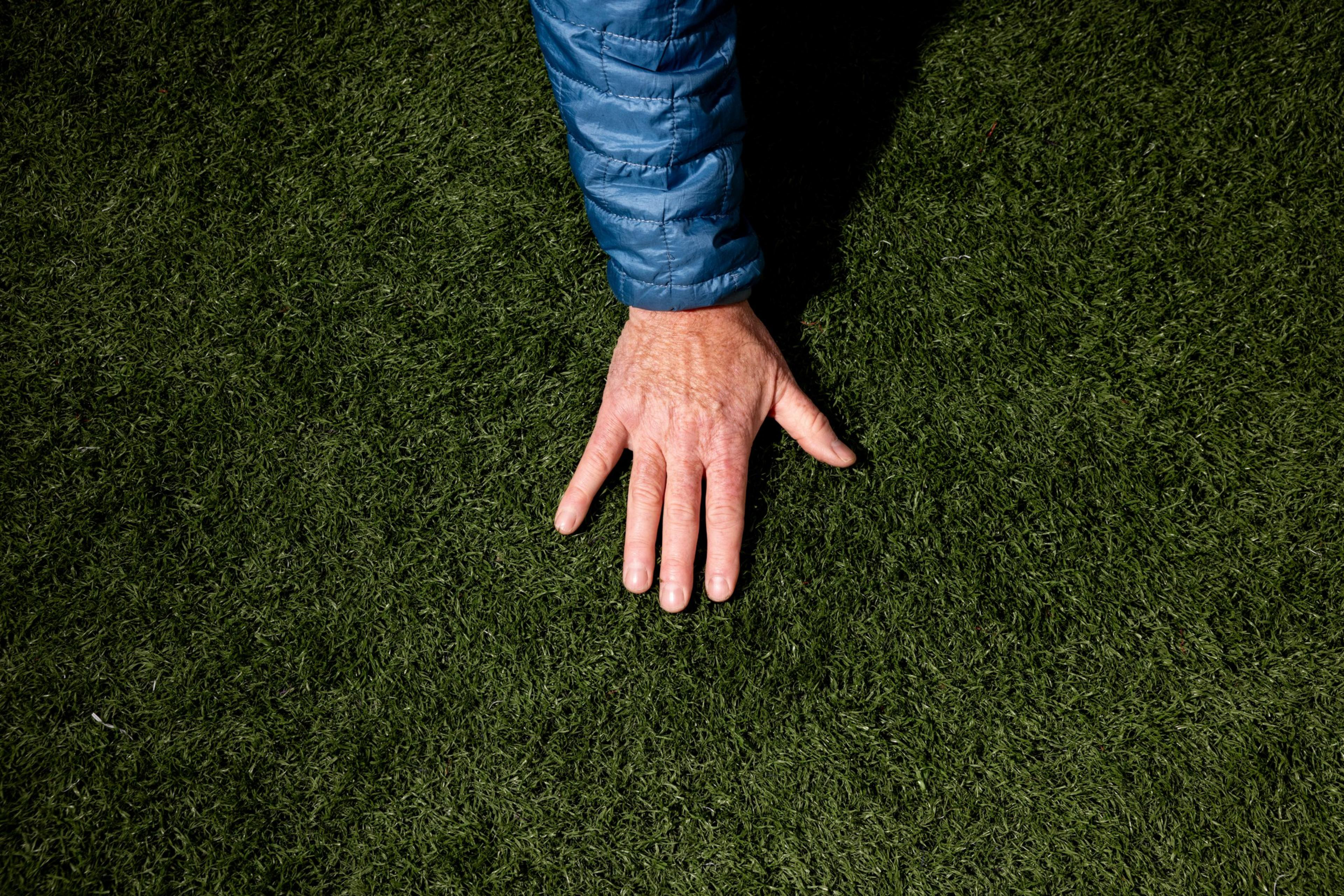 A hand in a blue jacket sleeve is placed on lush green grass, with fingers spread slightly apart.