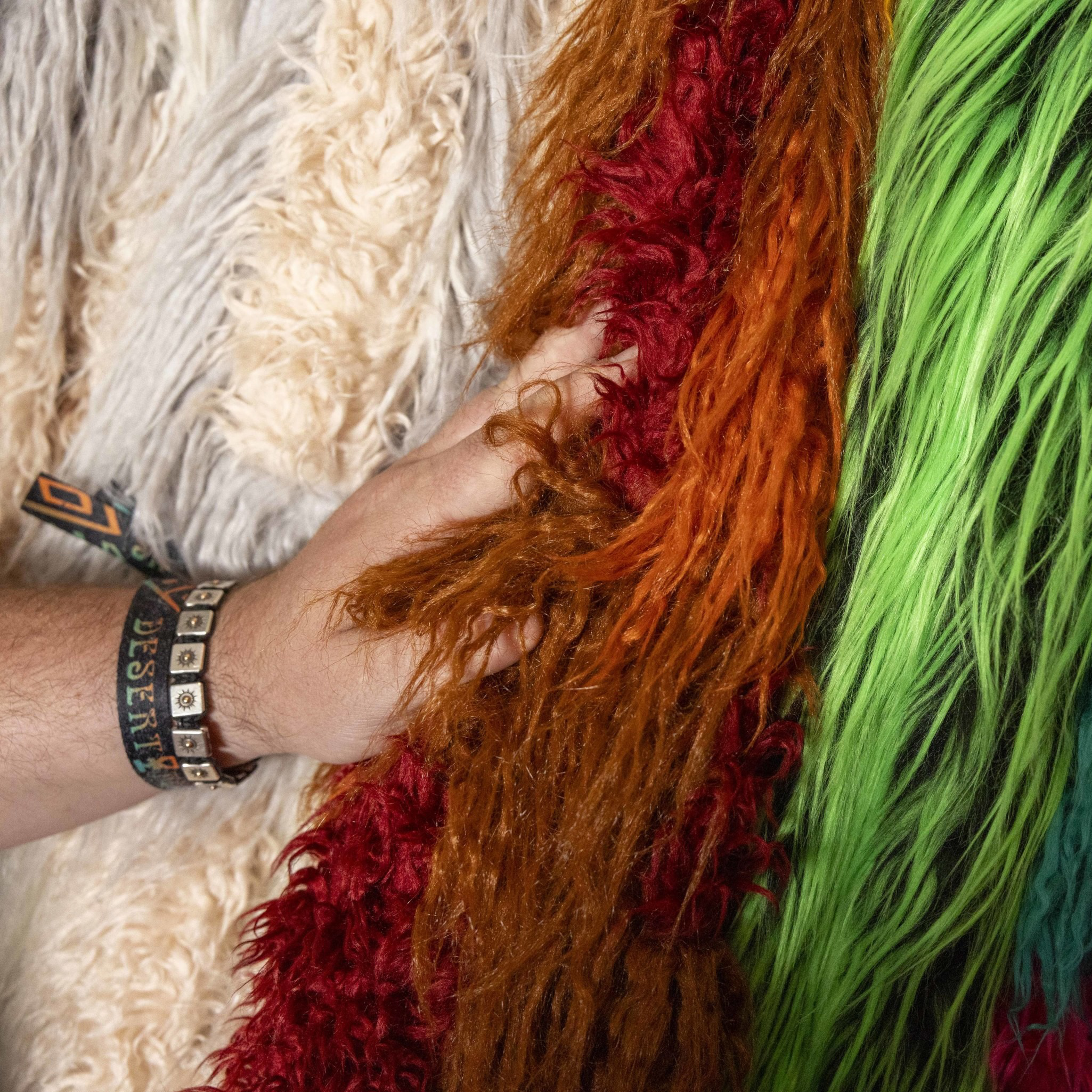 A hand is grasping fuzzy, textured fabrics in various vibrant colors, including green, red, orange, and cream, with the person wearing bracelets on their wrist.