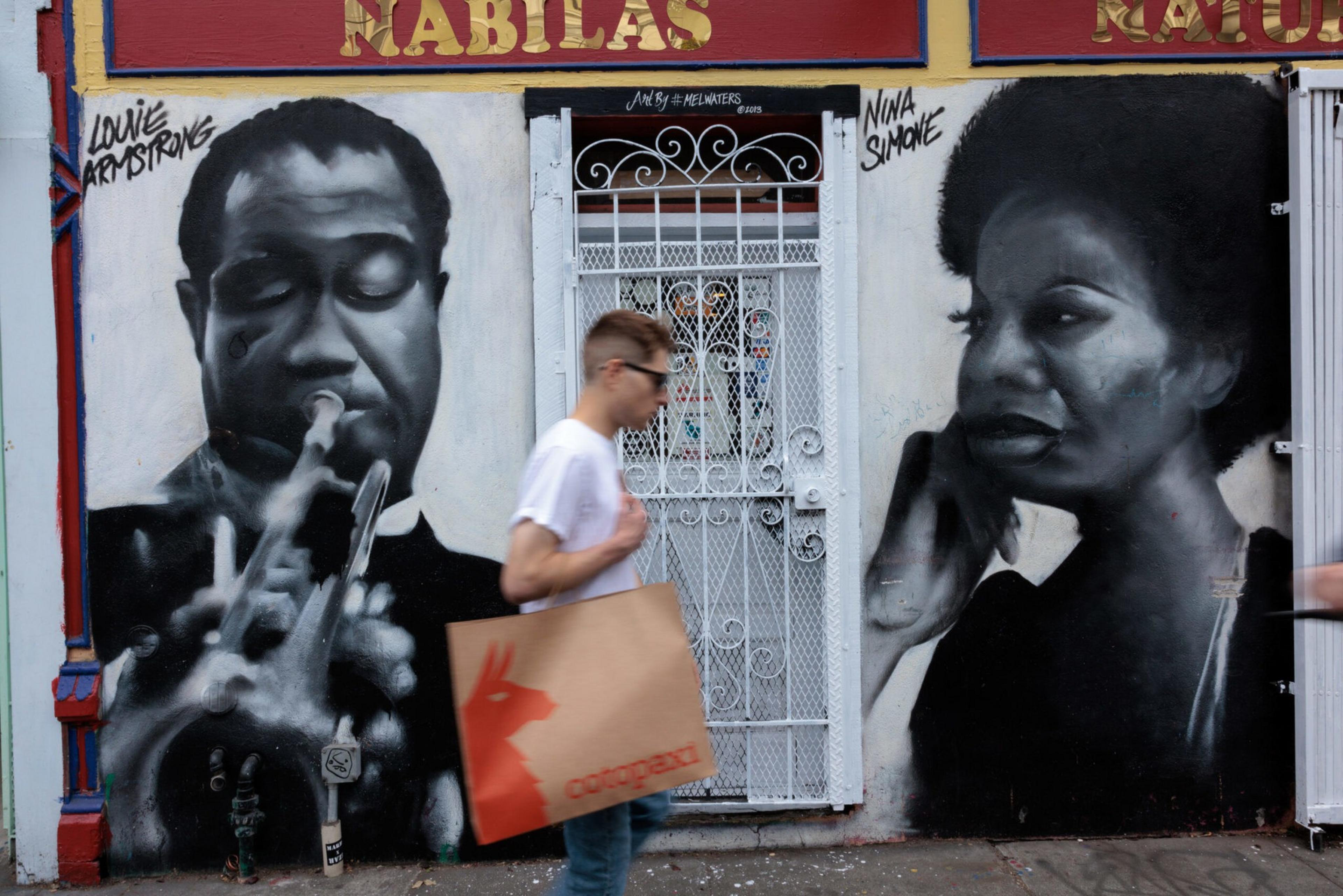 Black-and-white murals of Louis Armstrong playing trumpet and Nina Simone singing flank a white metal door, with a man carrying a shopping bag walking past.