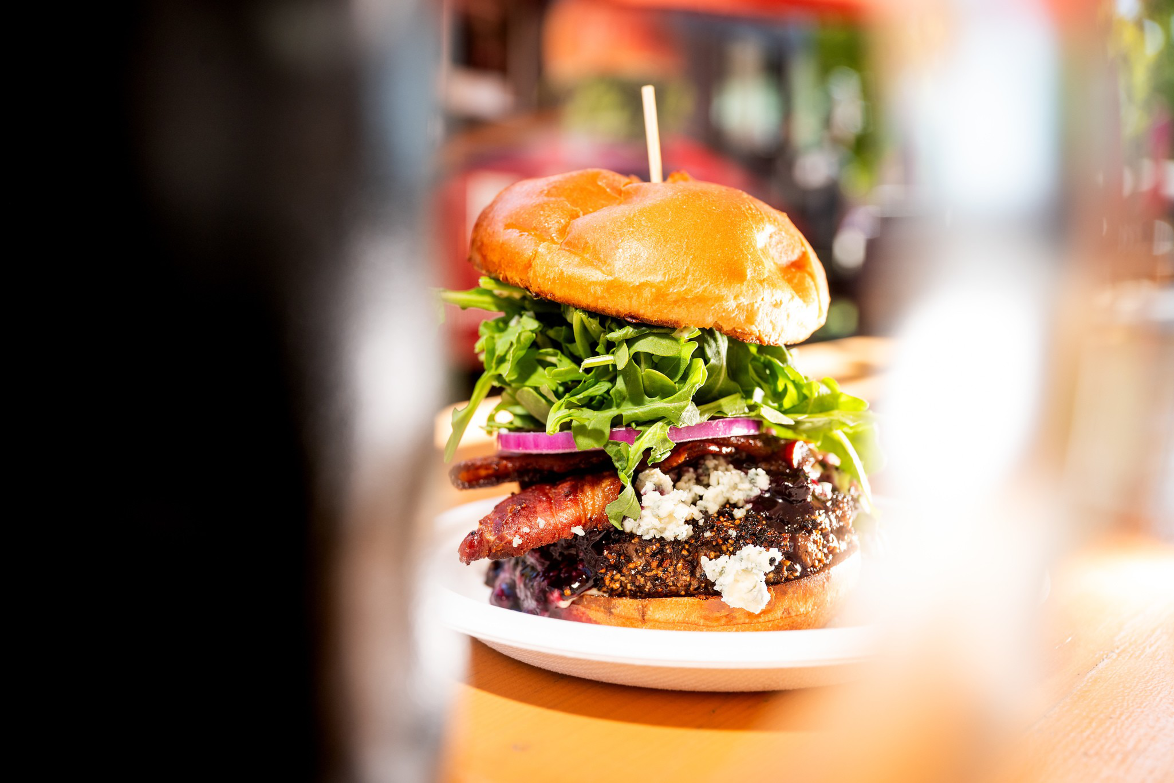 A burger with crispy bacon, crumbled cheese, red onion, a thick patty, and fresh leafy greens, served on a toasted bun with a toothpick on top.