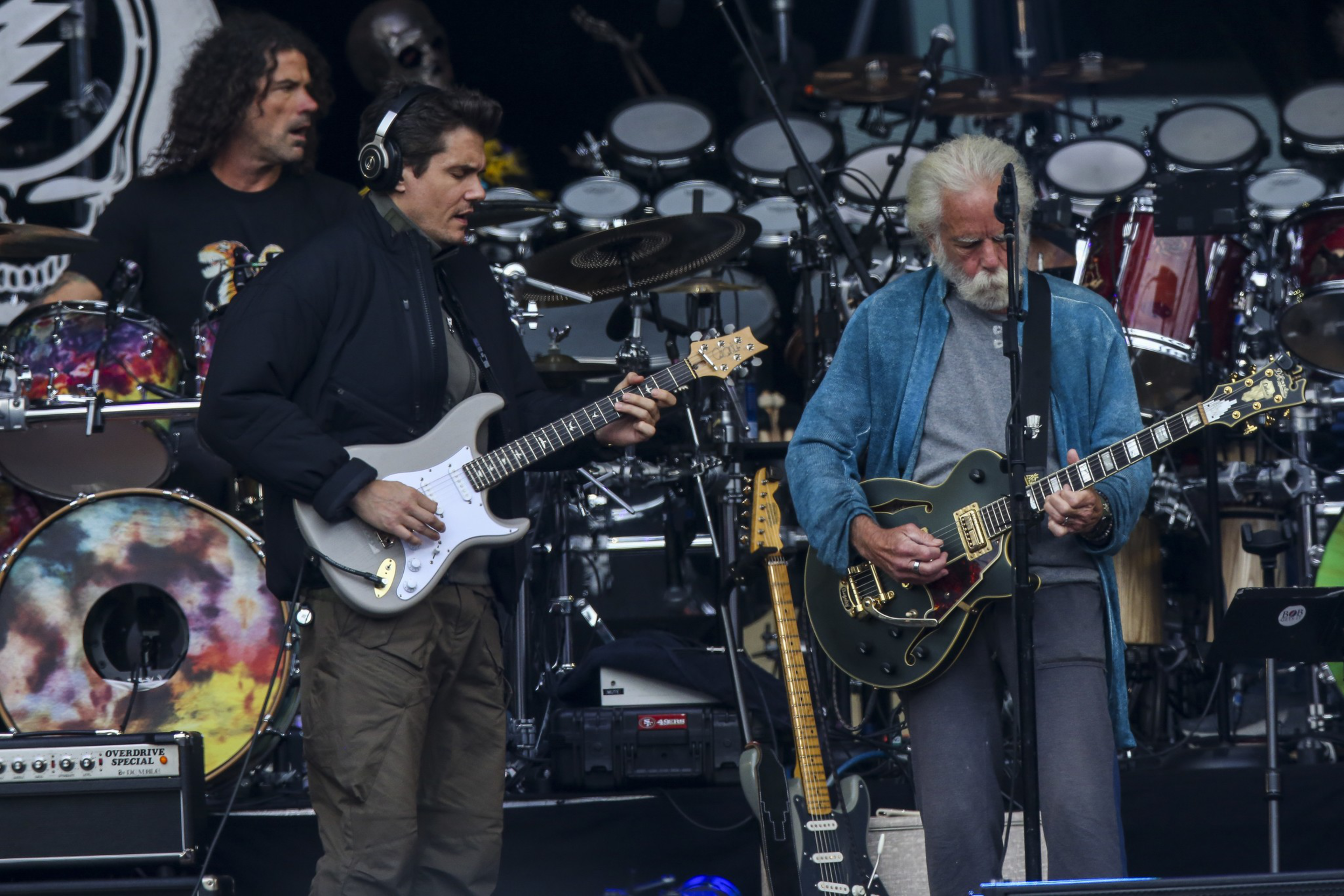 Two men are playing electric guitars on stage, surrounded by drums. One wears a dark jacket and the other a blue cardigan. Their faces show concentration.