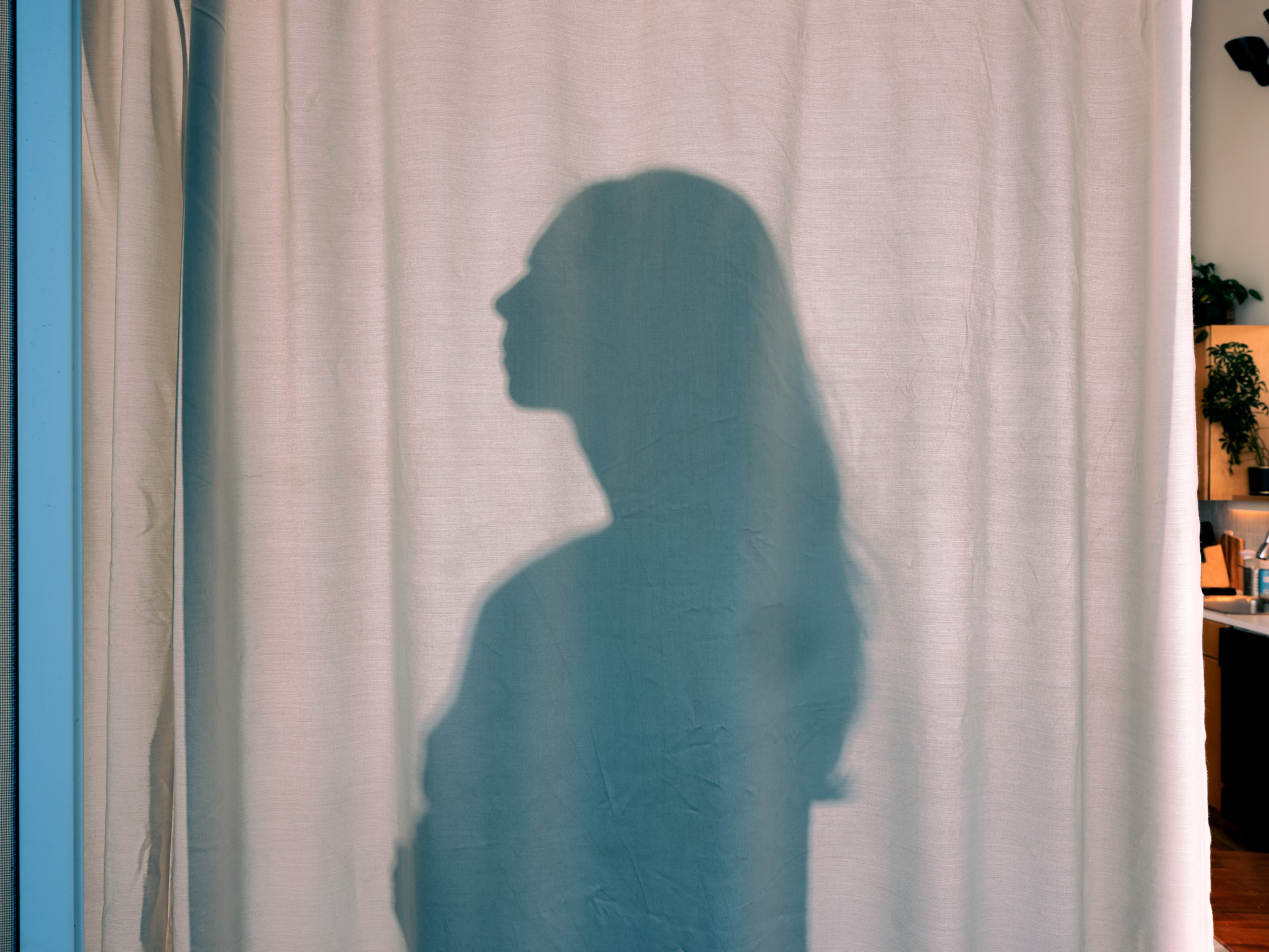 A silhouette of a person with long hair is visible behind a light-colored curtain, with a kitchen setting faintly seen on the right side.