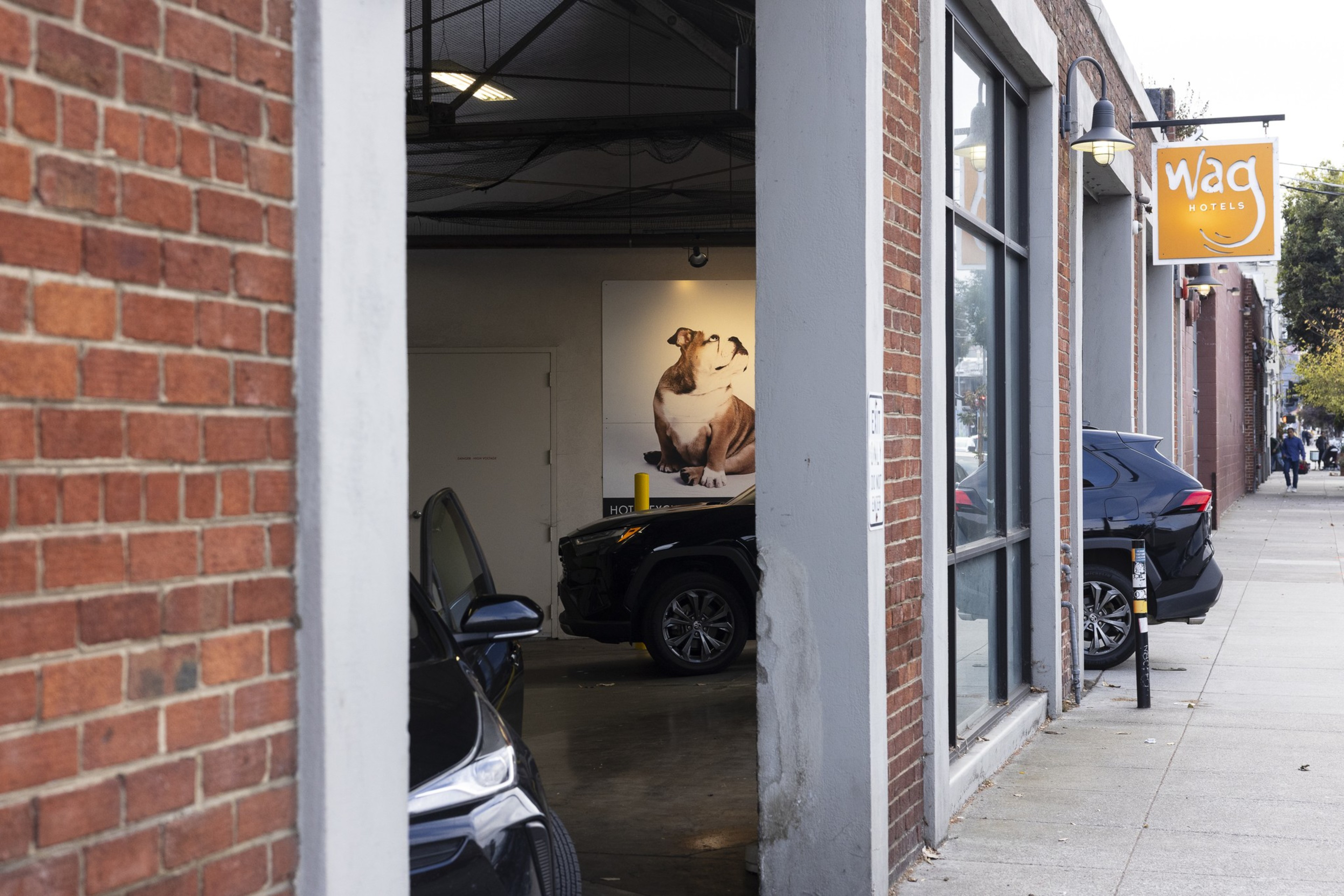 A brick building with a "wag hotels" sign shows a garage with two black cars and a large photo of a bulldog on the interior wall.