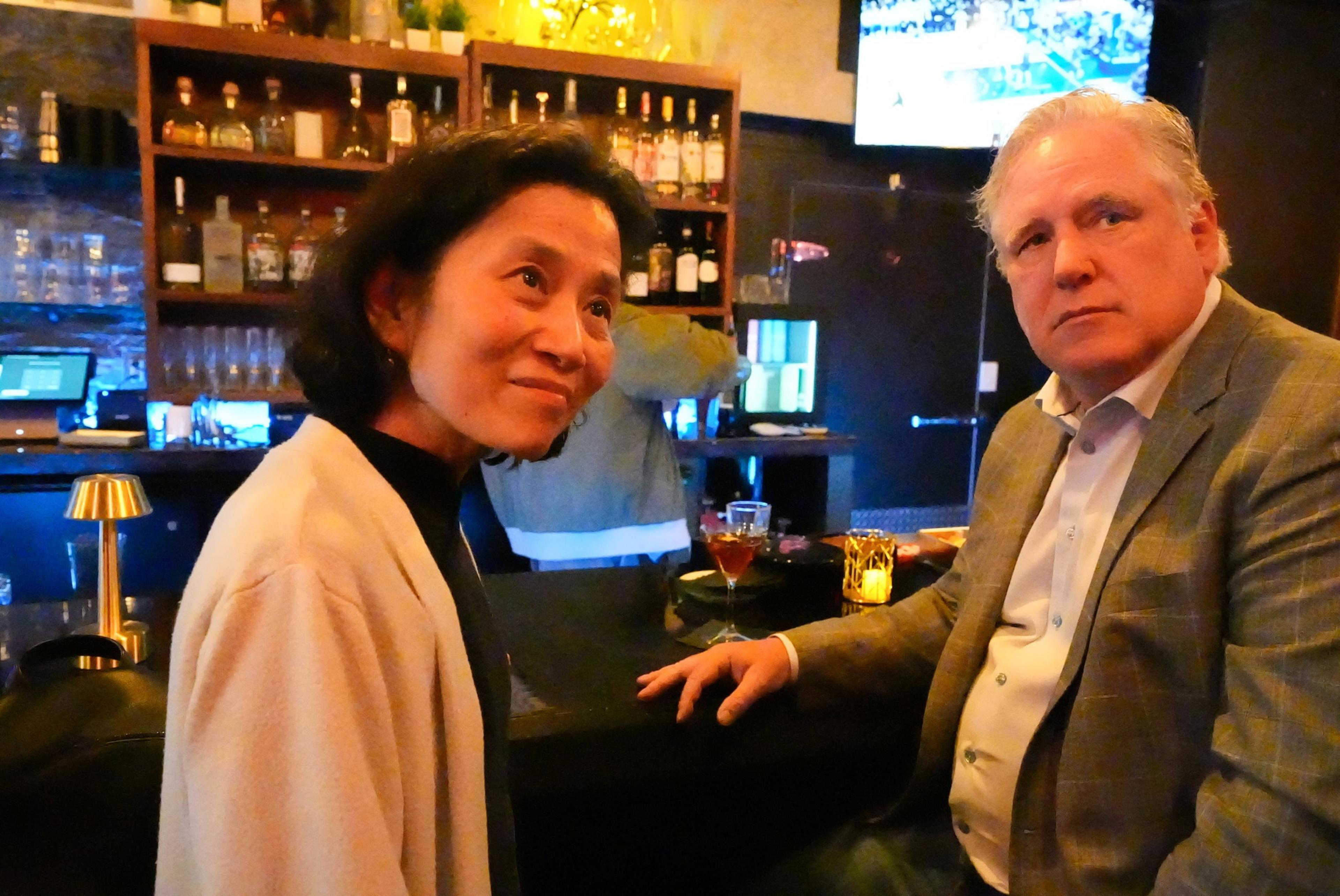 A man and woman sit at a dimly lit bar, facing the camera. Shelves with bottles and glassware fill the background, and a TV screen shows a sports game.