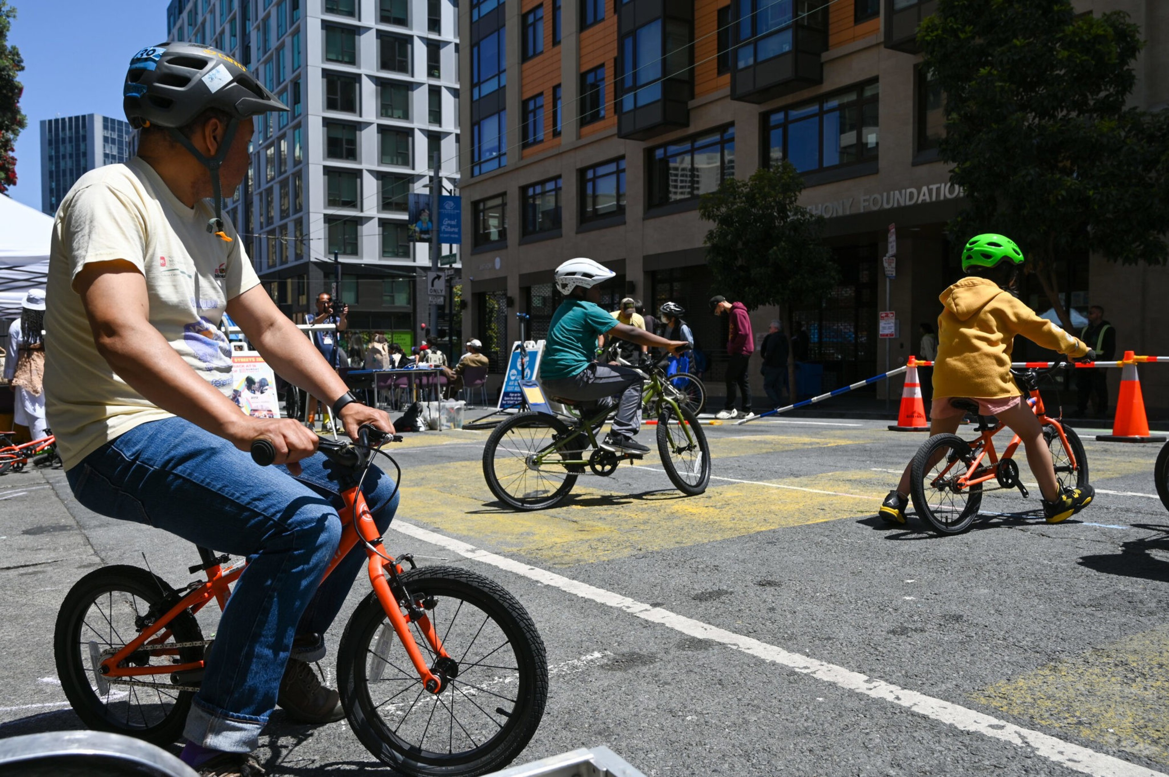 A group of people, including a child, are cycling on a city street with buildings and cones around.