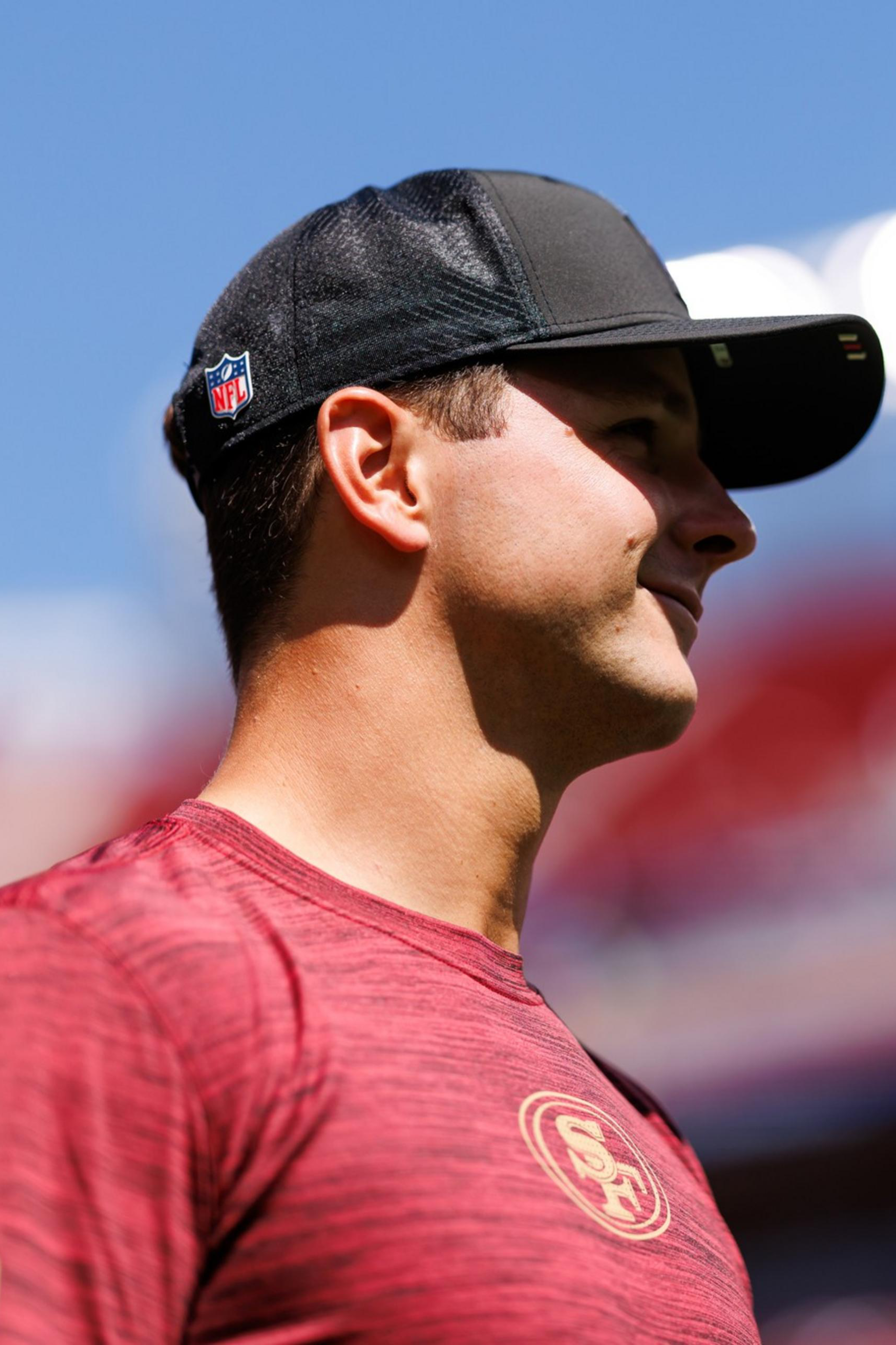 A man wearing a red San Francisco 49ers shirt and a black NFL cap stands, looking to the right against a bright, blurred stadium background.