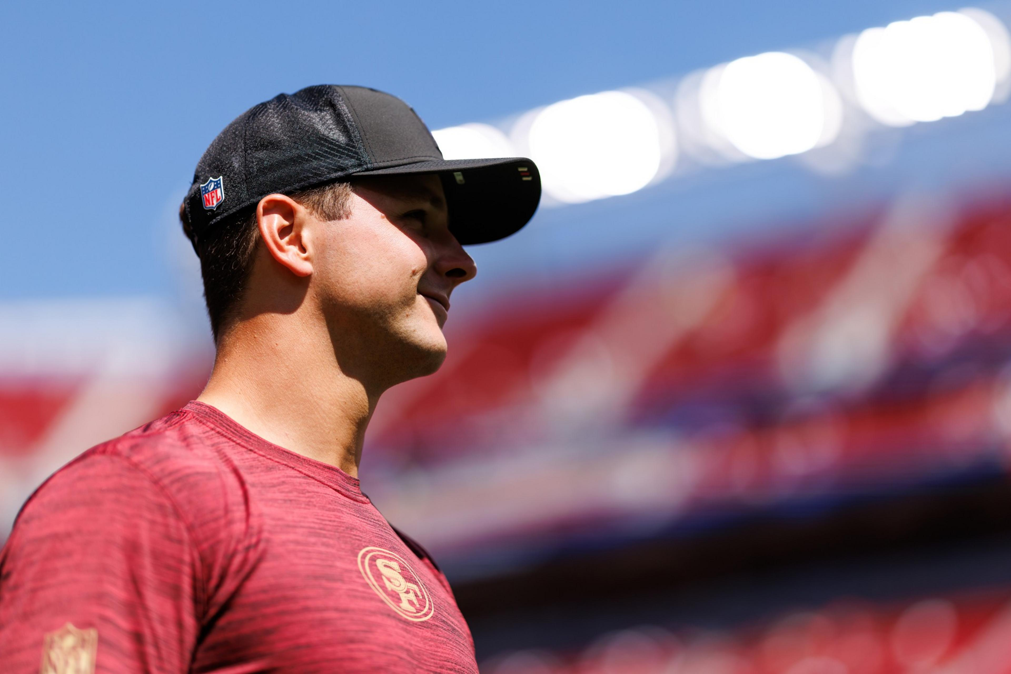 A man wearing a red San Francisco 49ers shirt and a black NFL cap stands, looking to the right against a bright, blurred stadium background.