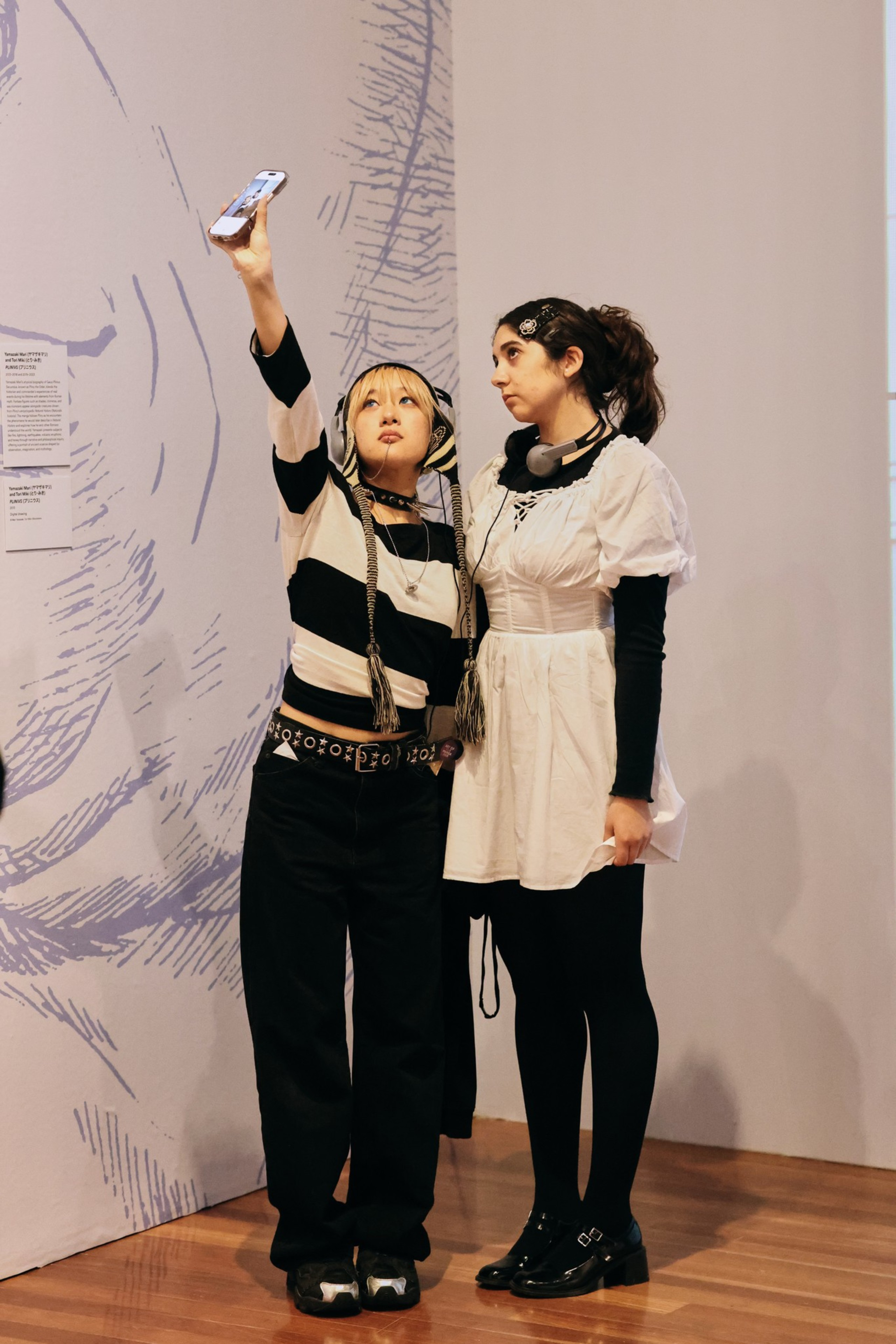 Two young women stand indoors, one wears a black and white striped top and takes a selfie, while the other in a white dress and black long sleeves looks away.