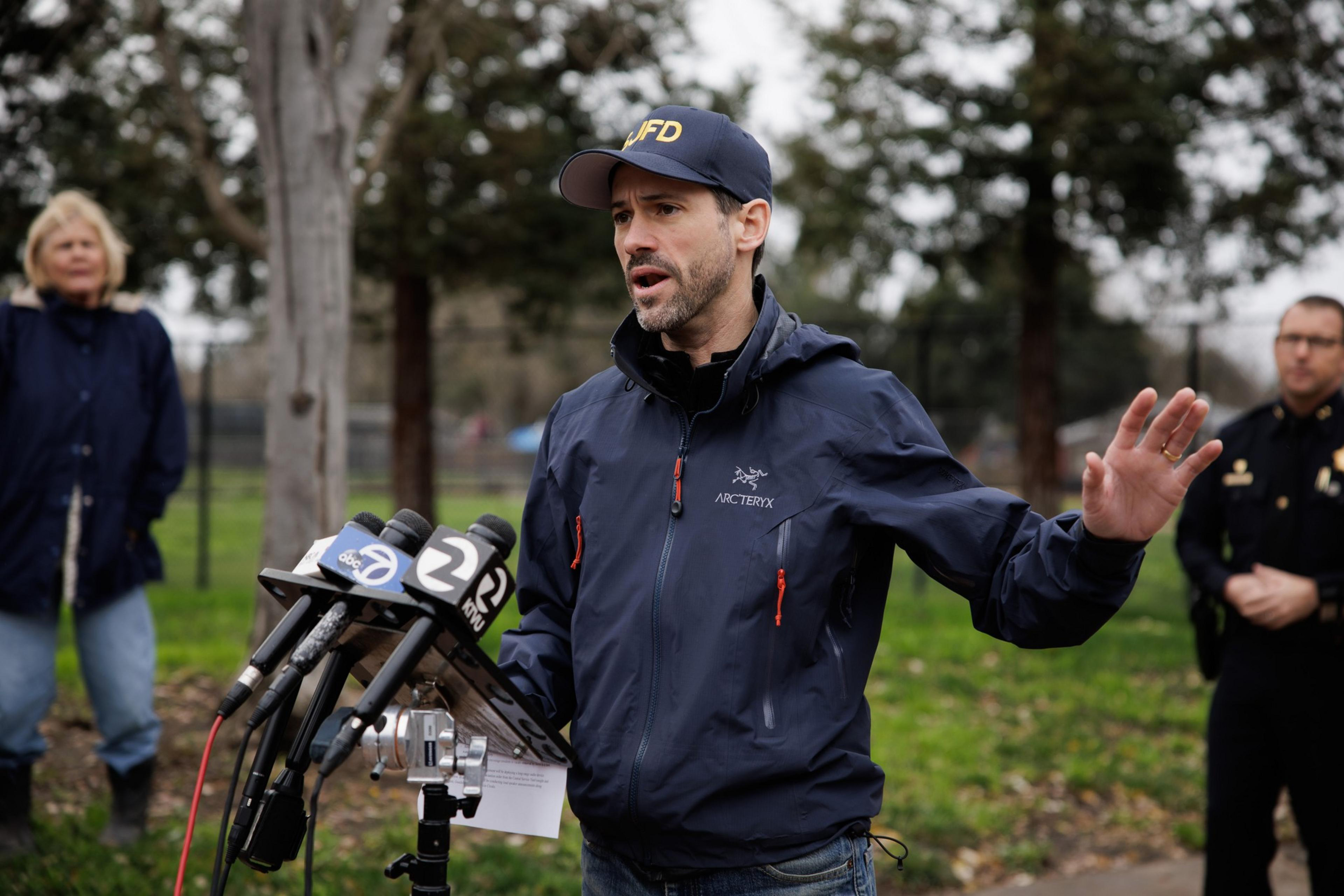 A man wearing a navy jacket and cap stands outdoors speaking at a microphone stand with news logos, gesturing with one hand while two people listen behind him.
