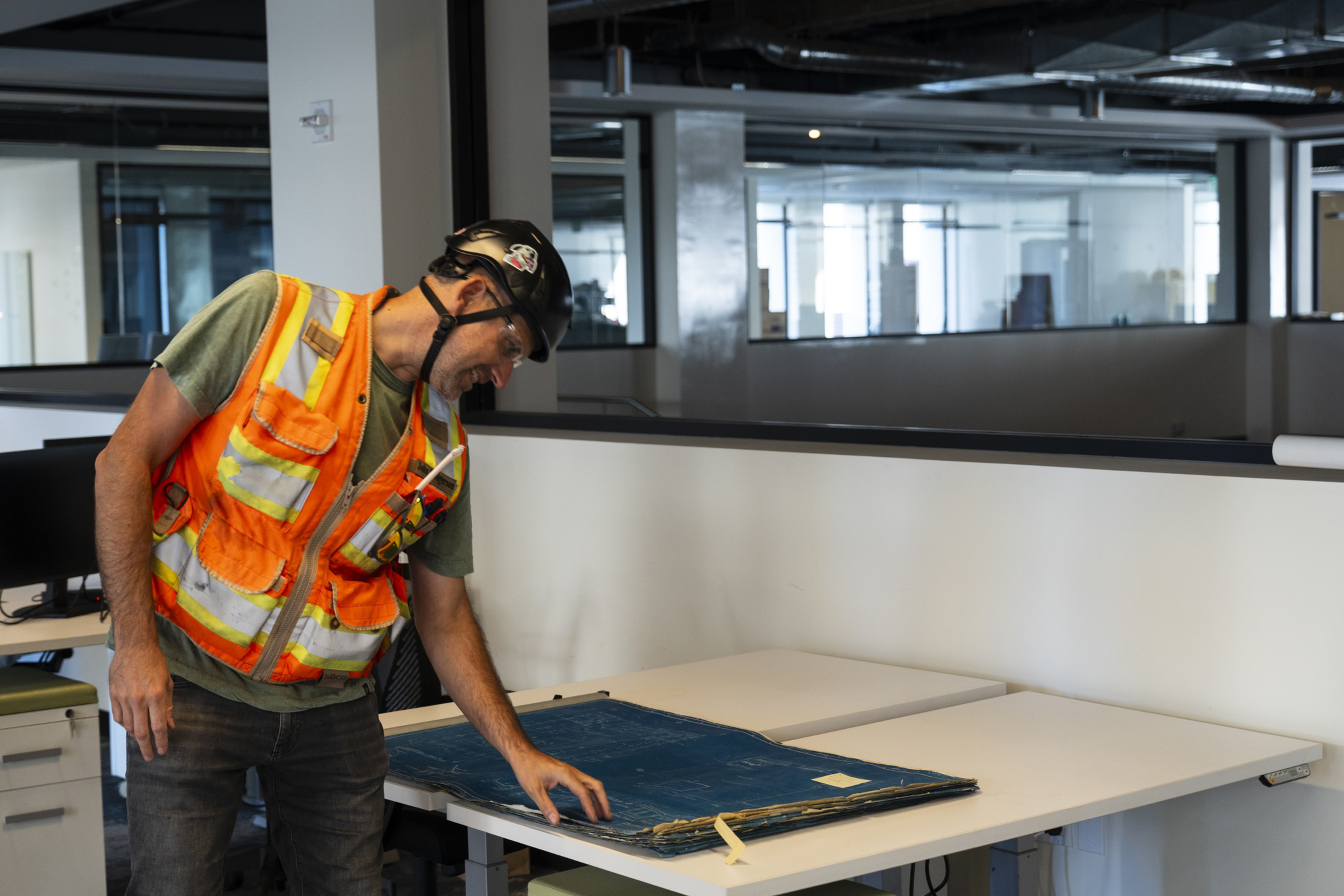 A construction worker in an orange safety vest and helmet examines blueprints spread out on an office desk.