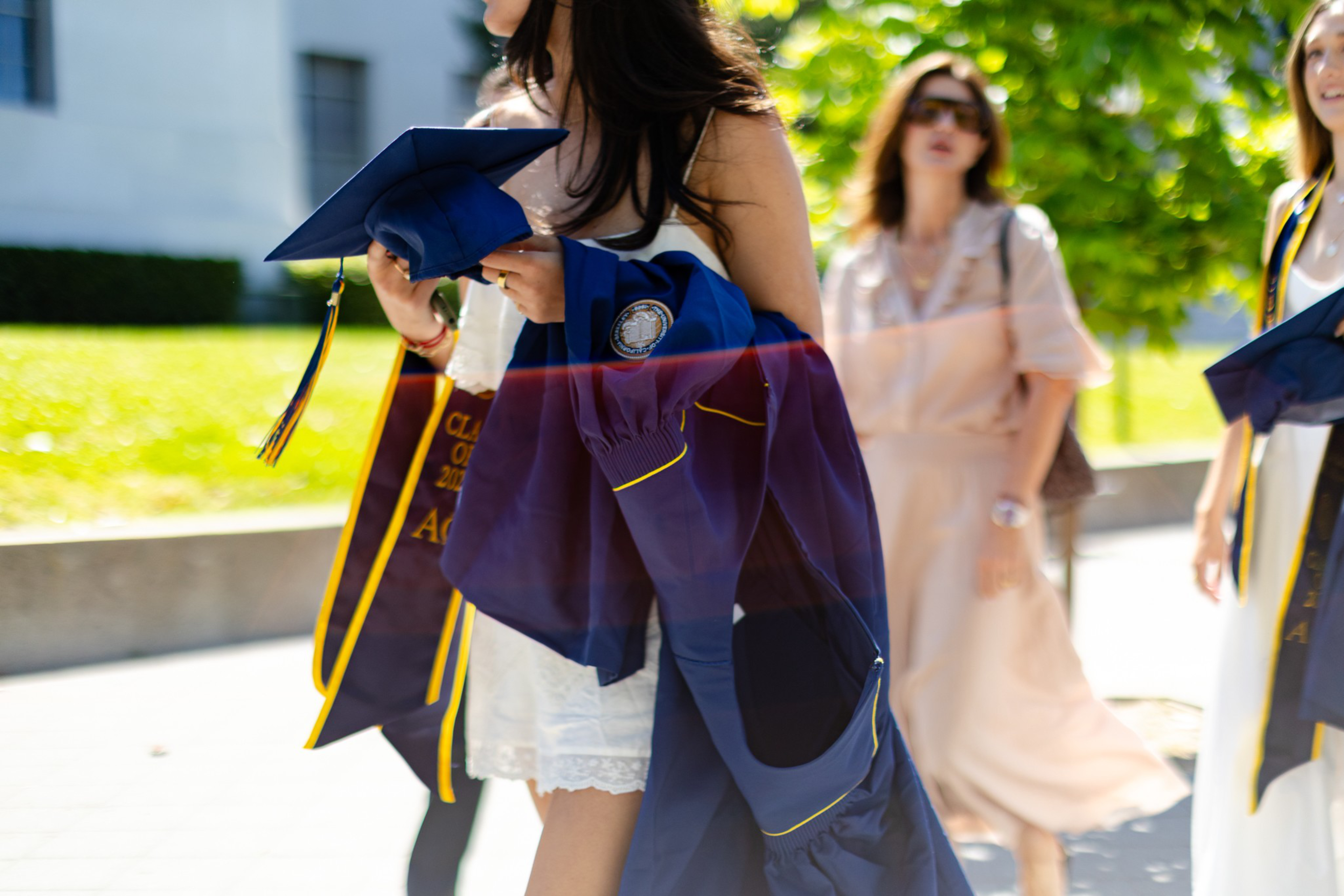 A person in a white dress holds a blue graduation cap and gown, with others and greenery blurred in the background on a sunny day.