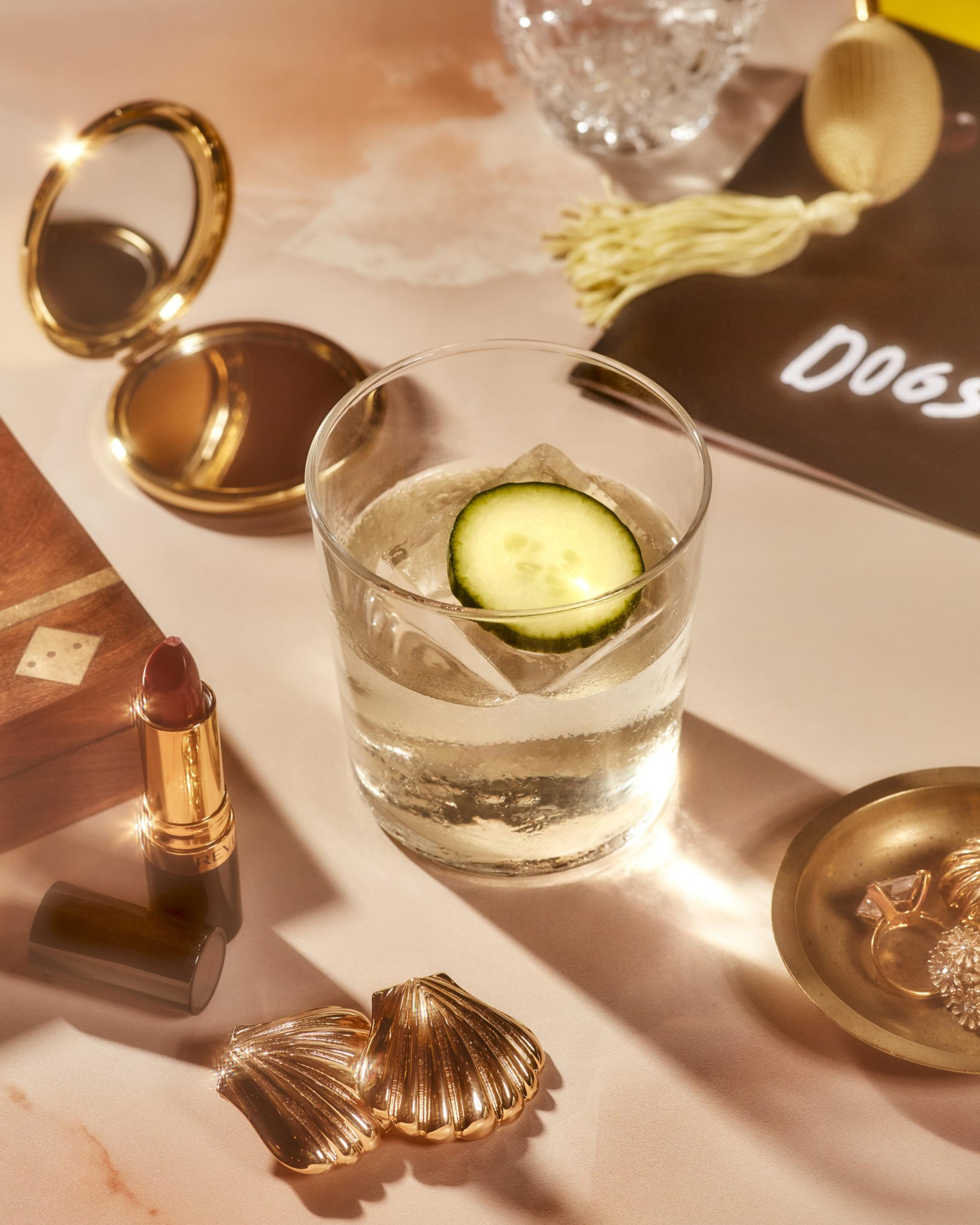 A cocktail with a cucumber slice sits on a table with a gold compact, red lipstick, jewelry, a perfume bottle, and a crystal glass. The scene is elegant and refined.