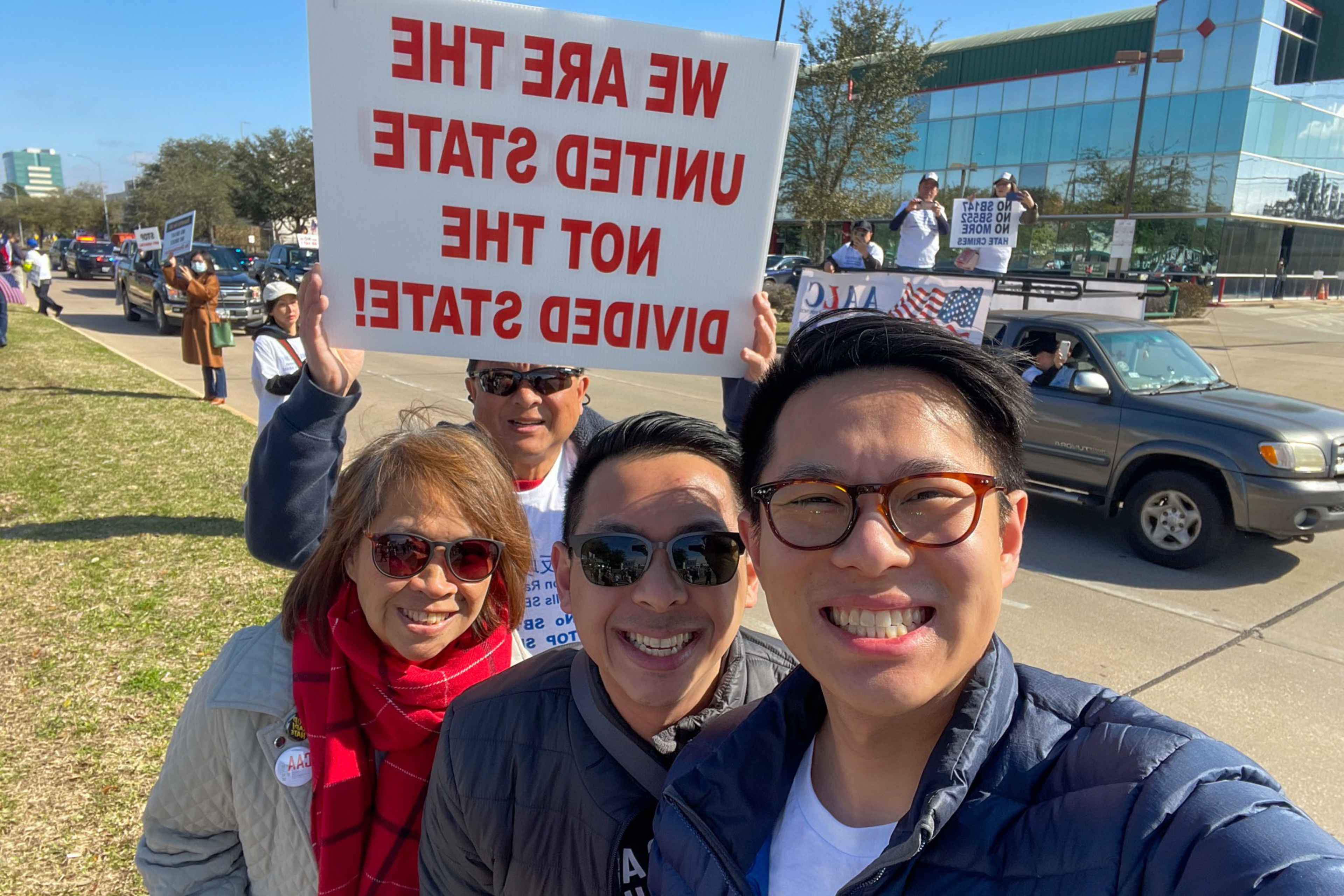 A group of people smiling at the camera during a protest, with one holding a sign reading, "WE ARE THE UNITED STATE NOT THE DIVIDED STATE!"