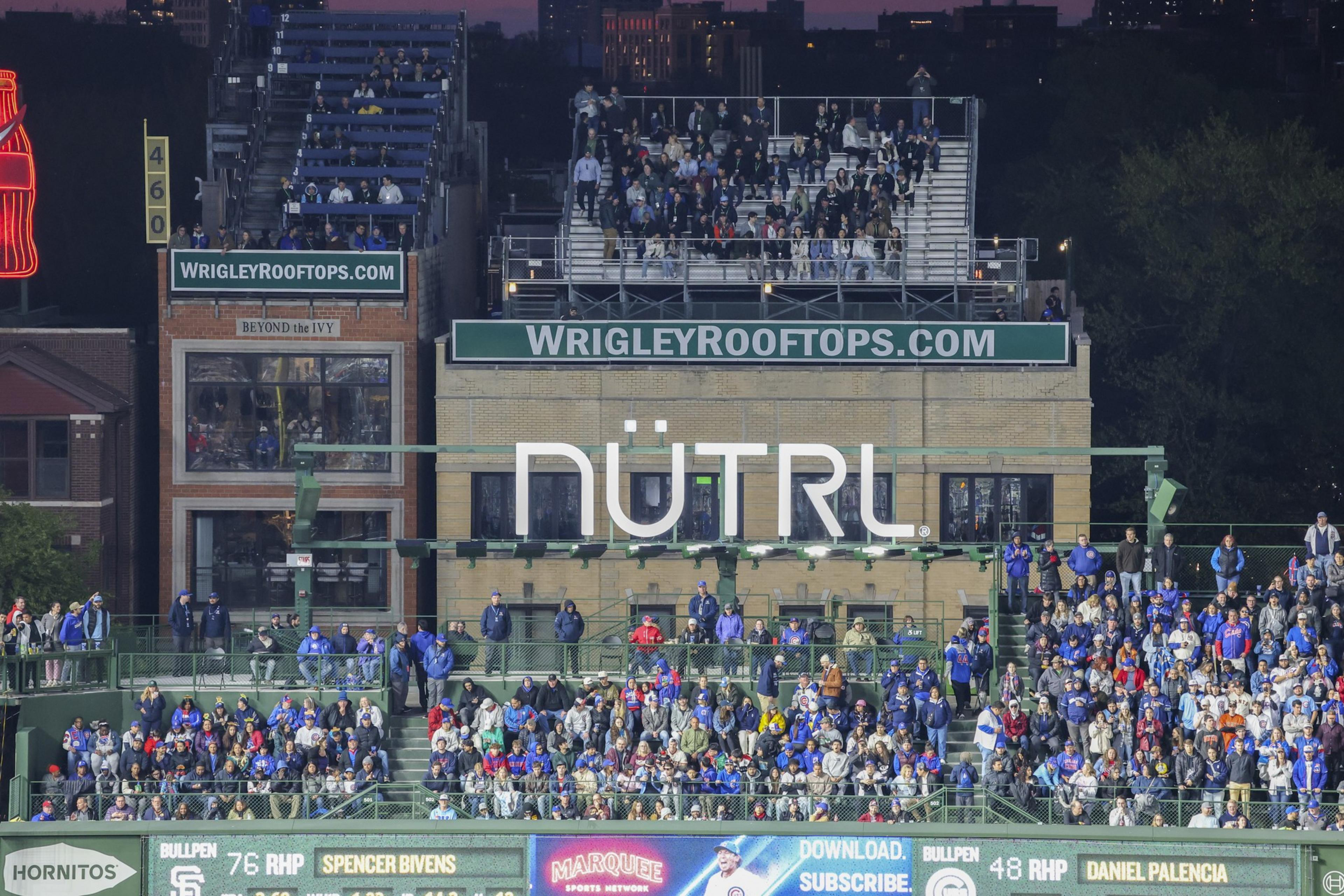 The image shows a crowded seating area above a building with a large &quot;NÜTRL&quot; sign. People are gathered on multiple levels, overlooking a sports venue.