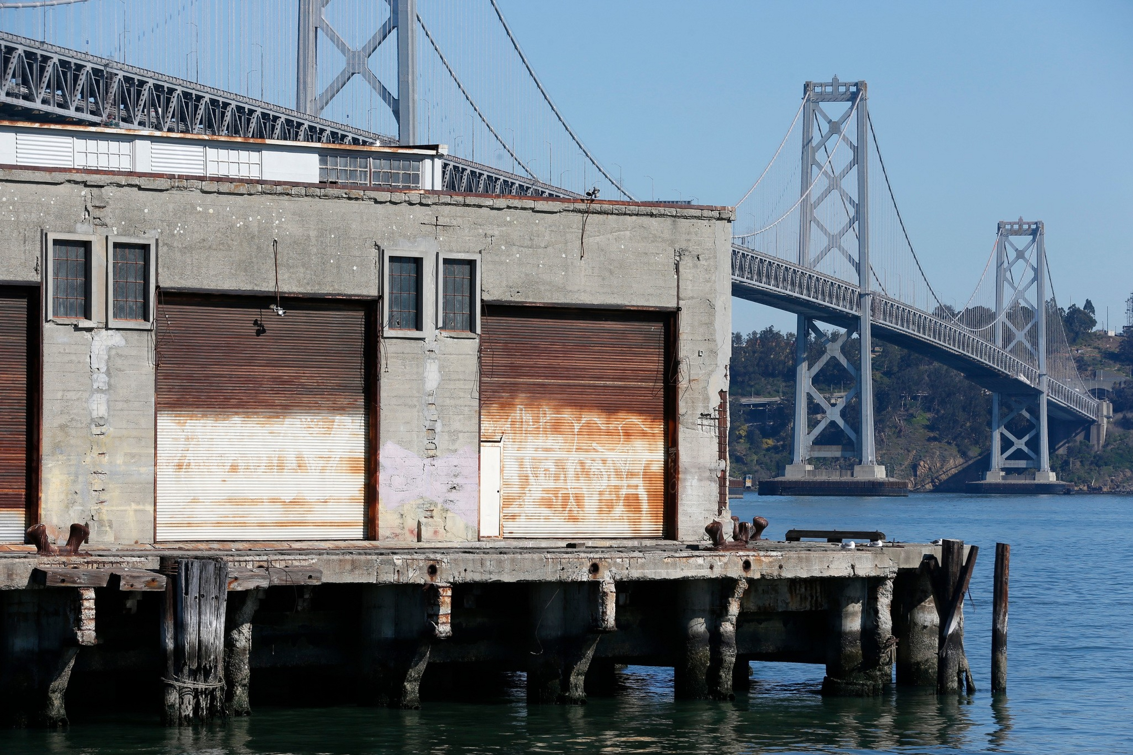 A weathered concrete pier building with rusted metal doors sits over calm water, with a large suspension bridge spanning across a clear blue sky in the background.