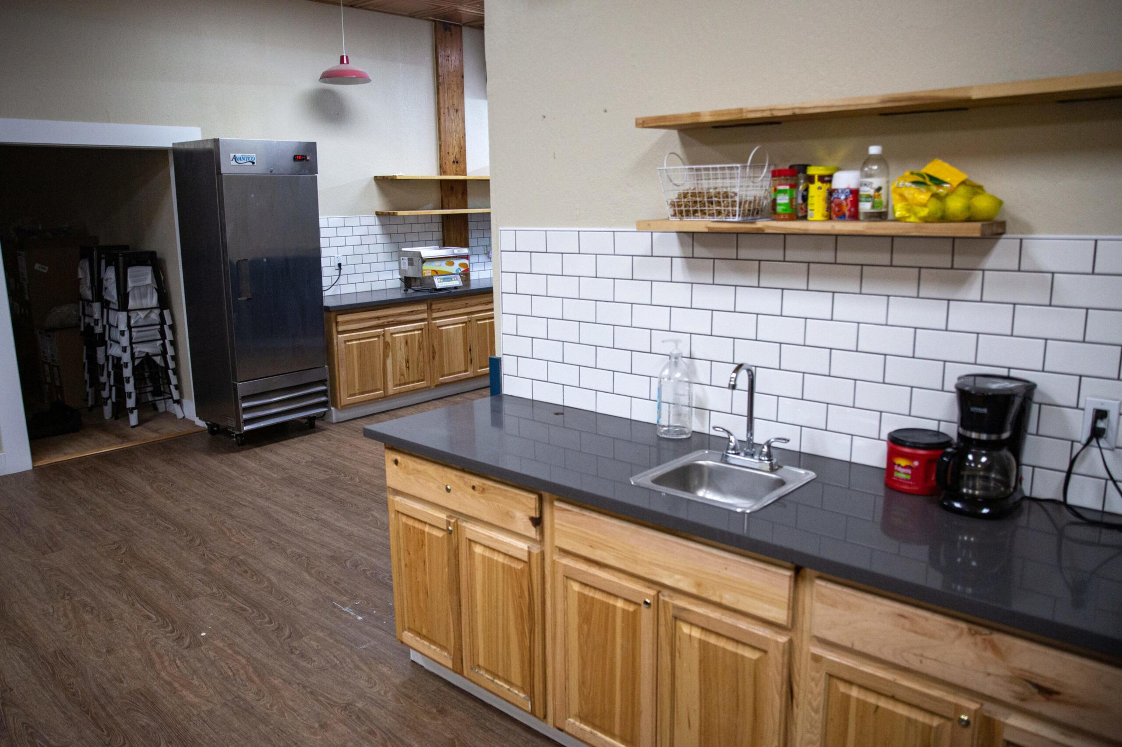 A kitchen with light wood cabinets, black countertops, a small sink, a coffee maker, a refrigerator, shelves holding jars, a basket, a bottle, and some lemons.