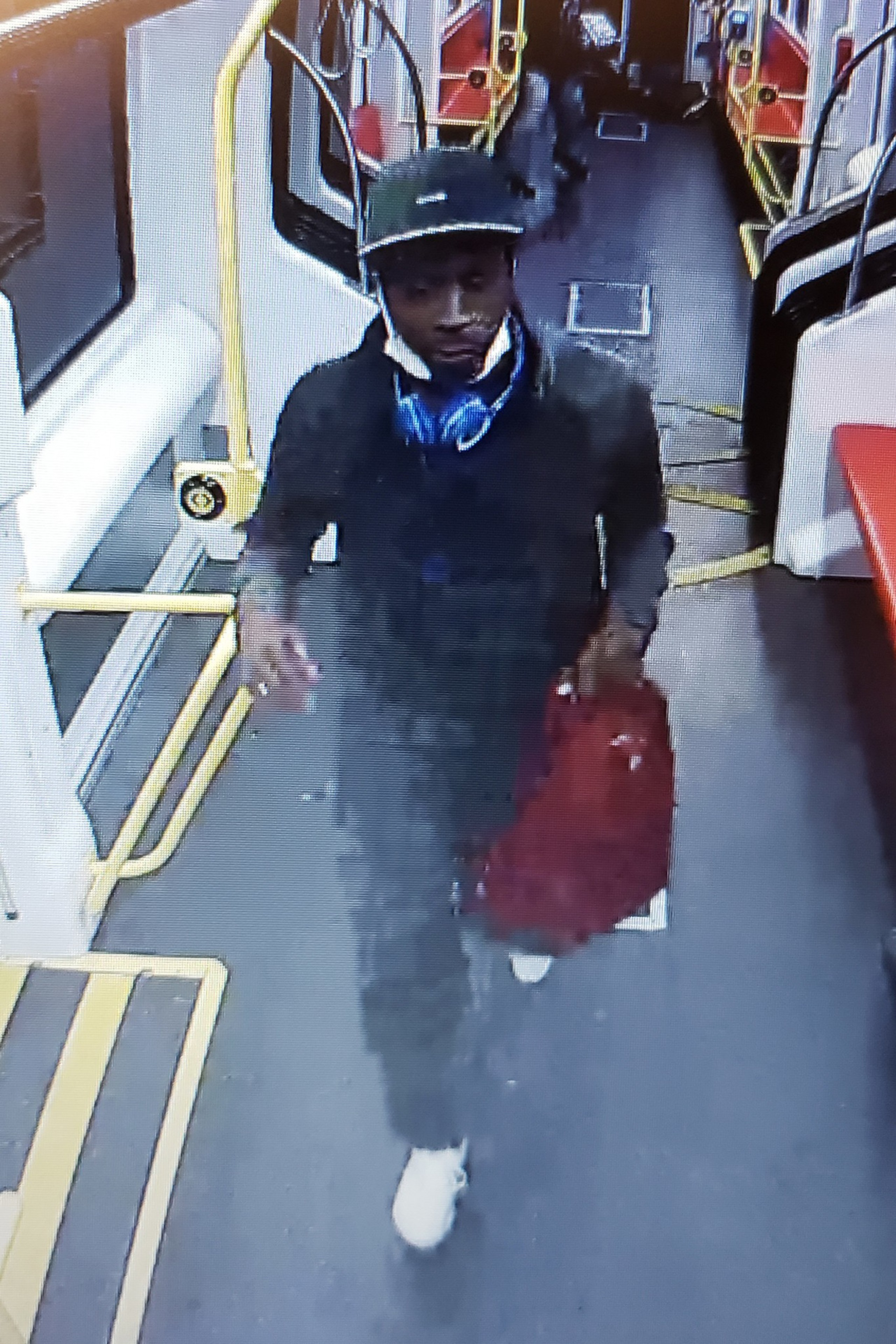 A man wearing a dark jacket and cap walks inside a transit vehicle, carrying a red bag and blue headphones around his neck.