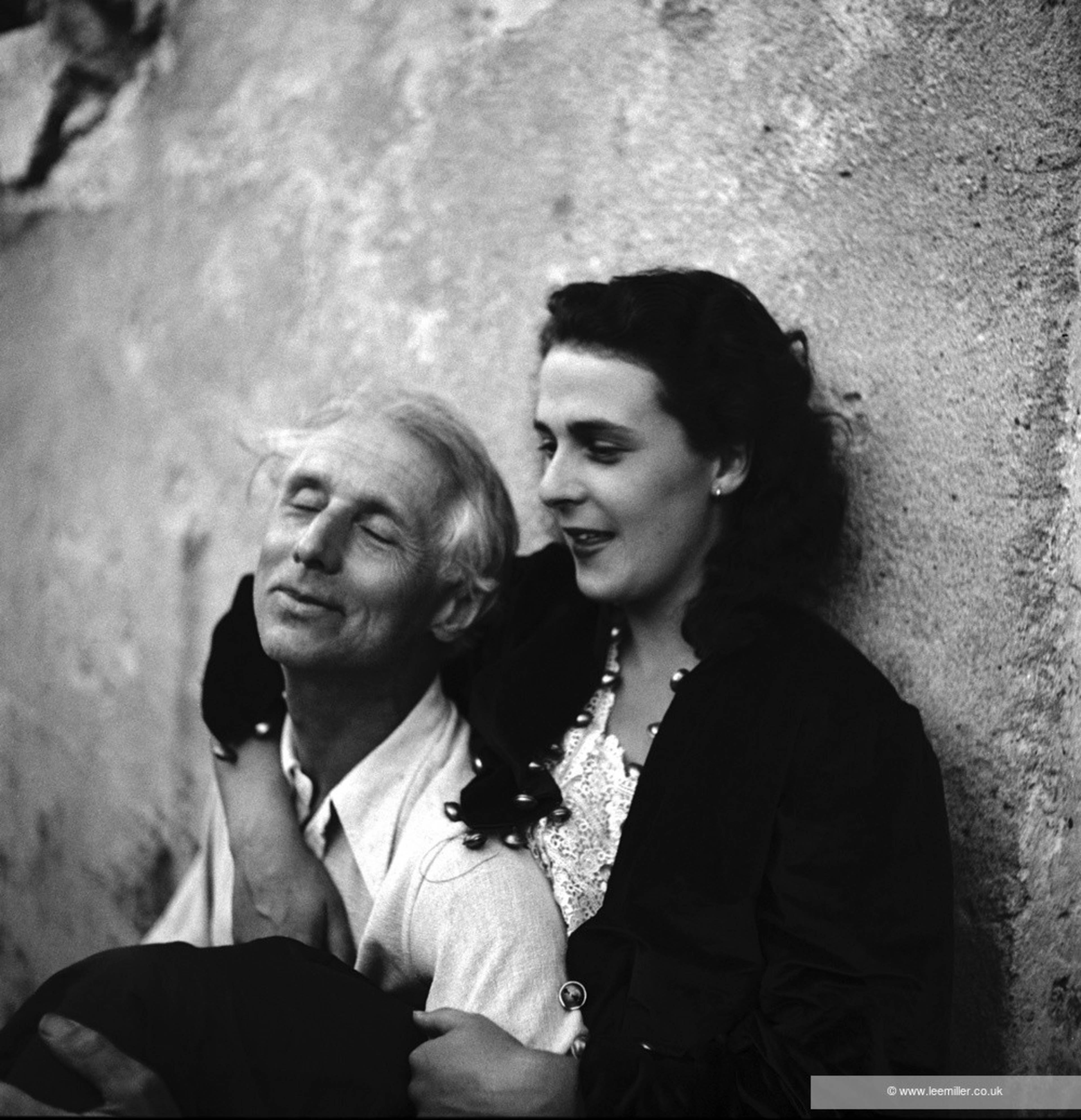 A man and woman are sitting closely together against a textured wall. The man has his eyes closed and wears a light shirt, while the woman, dressed in dark, smiles gently.