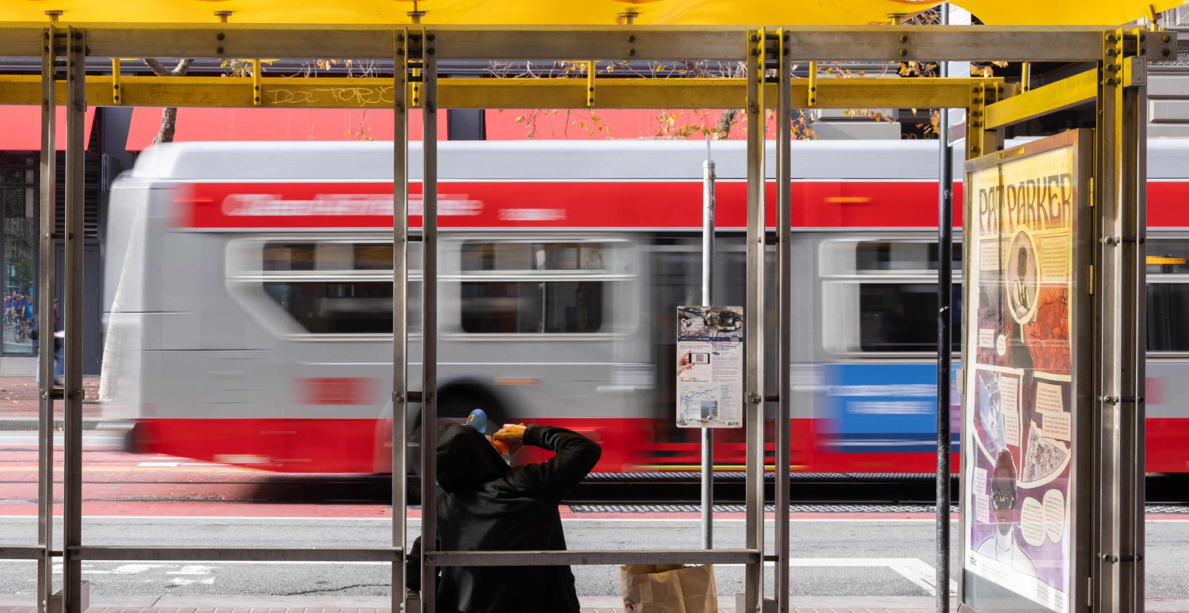 A person wearing a hoodie and face mask sits inside a yellow bus stop shelter while a red and white bus speeds by outside.
