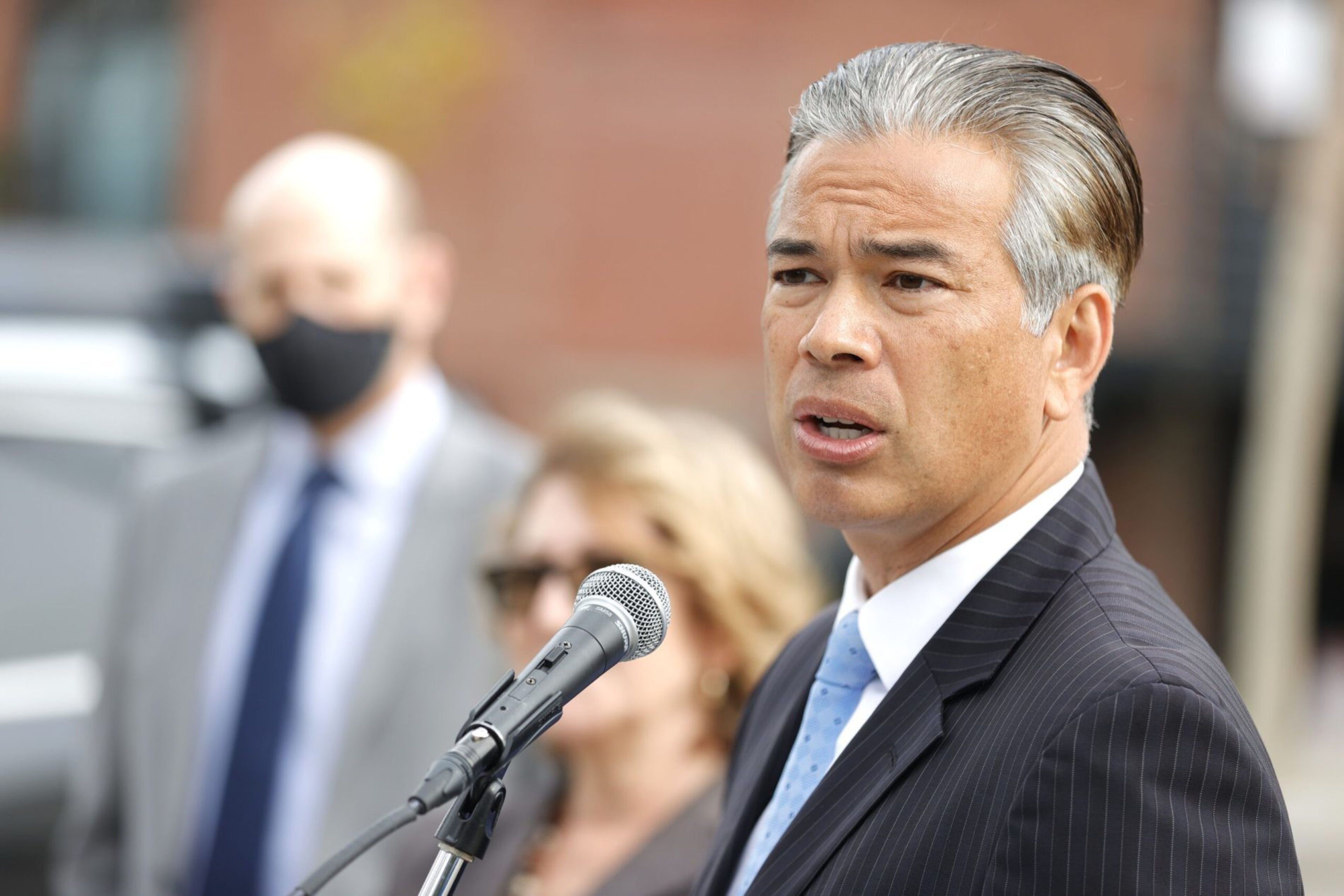 A man in a pinstripe suit and light blue tie speaks into a microphone, with two blurred people standing behind him outdoors.